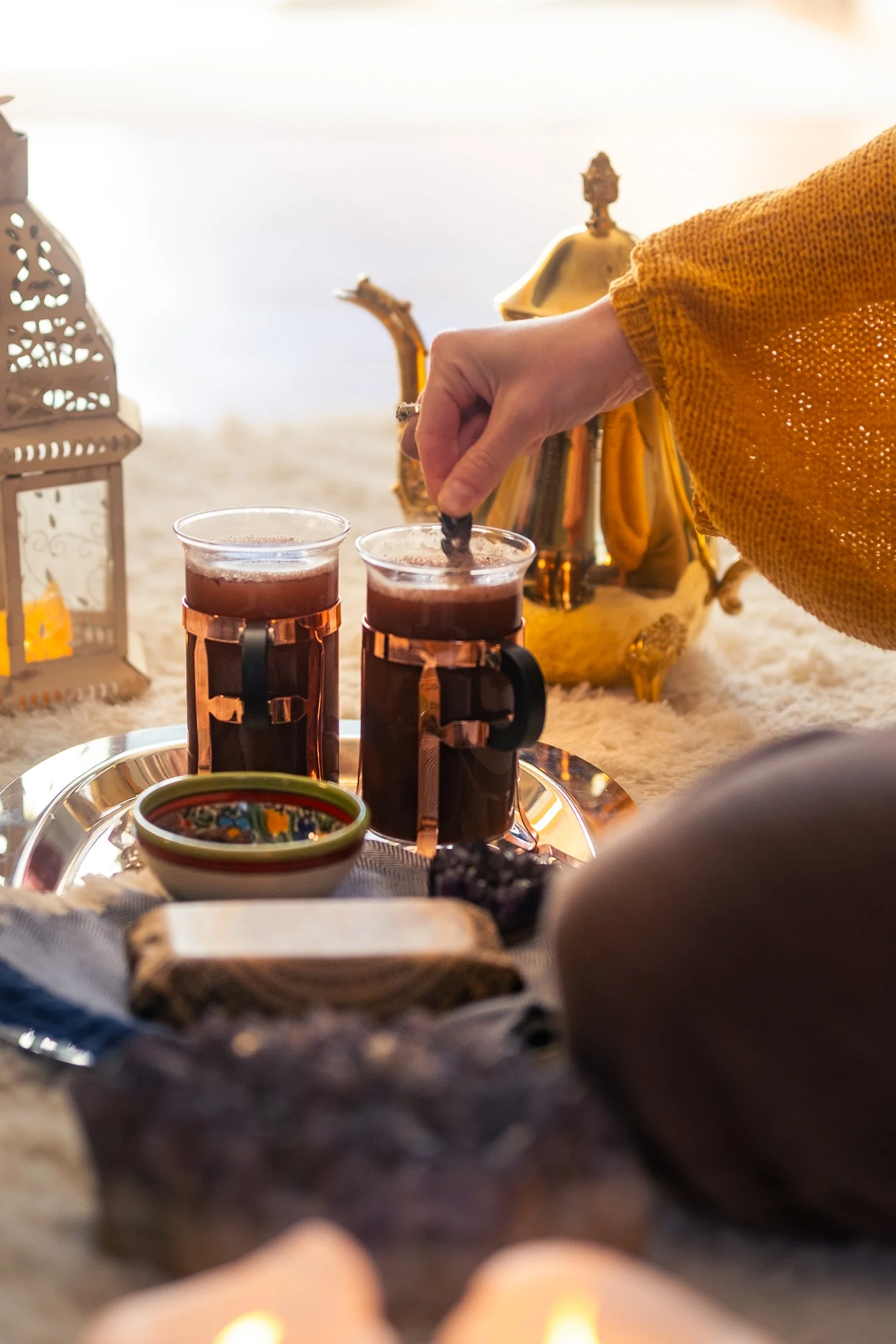 A person in a yellow sweater is stirring a glass of dark liquid, possibly tea, on a tray. There are two glasses filled with the same dark beverage, a decorative bowl, candles, and other objects on the tray, and a lantern to the left. The setting appears to be indoors with soft, natural lighting.