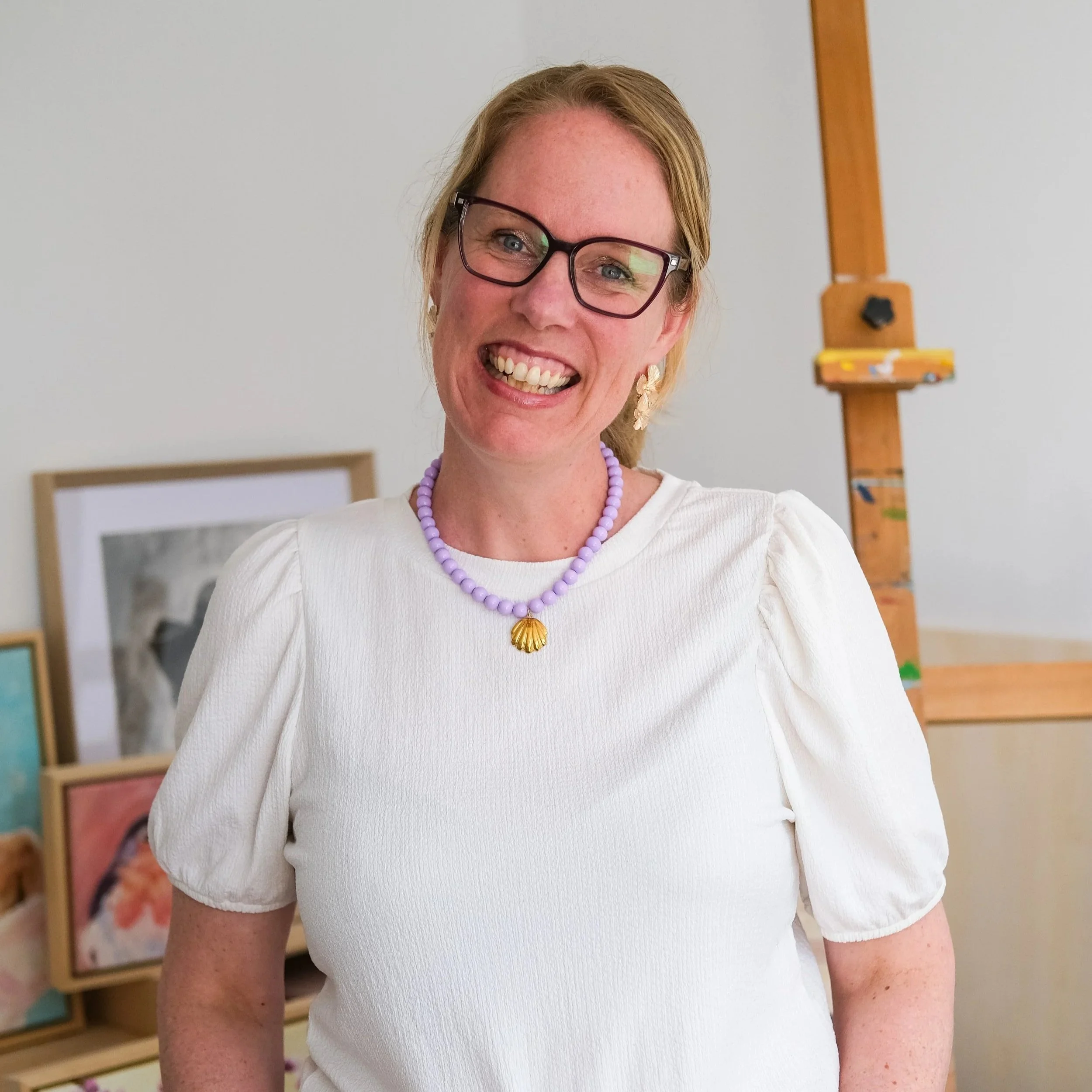 Close-up van een lachende vrouw met blauwe ogen en bruin haar, draag een paar zwart-randbrillen, een witte jurk, en een paarse parelketting en een gouden schelp-hanger, staand in een kamer met witte muren en een schilderijlijst op de achtergrond.