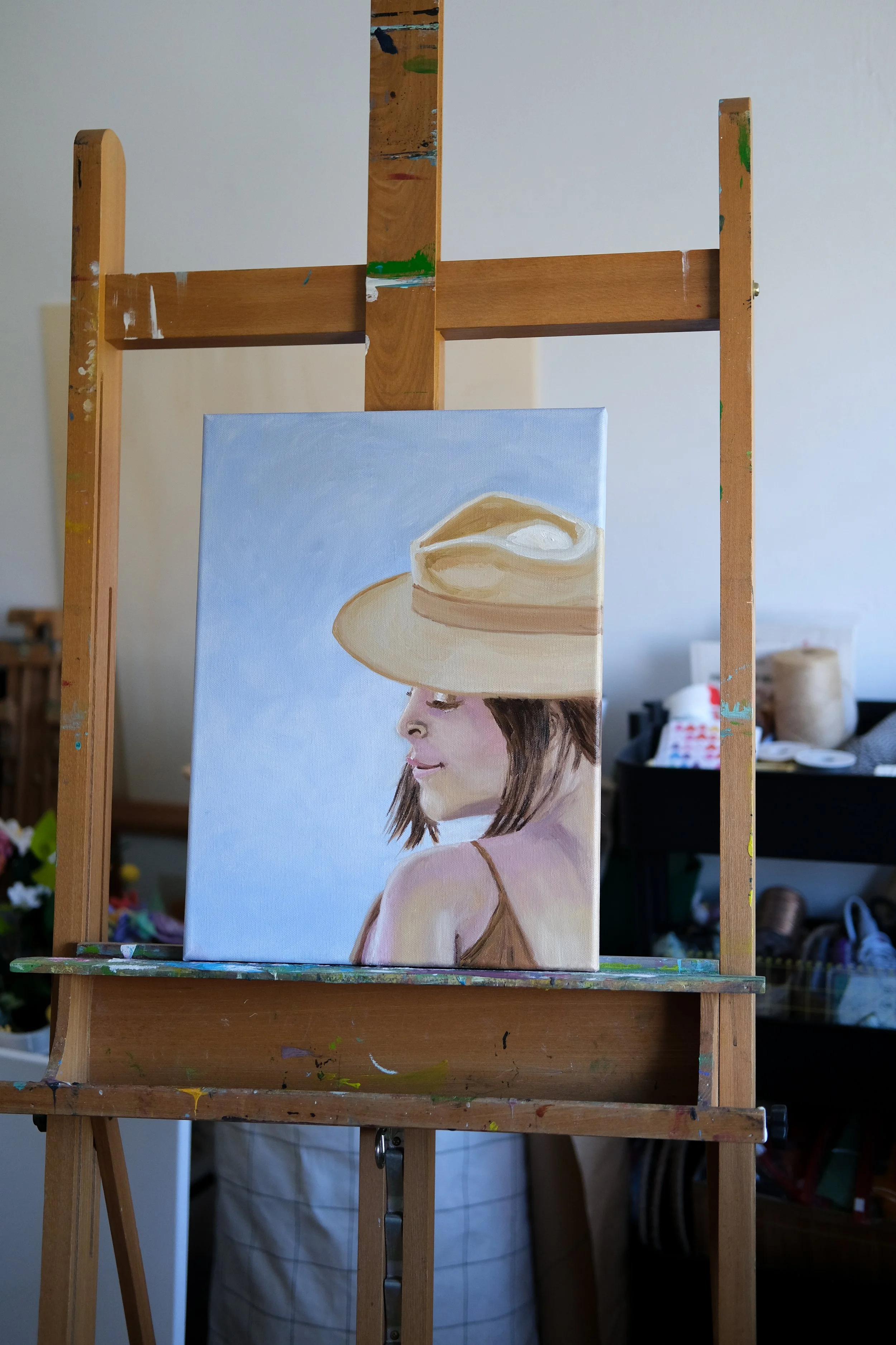 Schilderij van een jonge vrouw met een hoed, gemaakt in een schilderatelier met een houten ezel en schildermateriaal op de achtergrond.