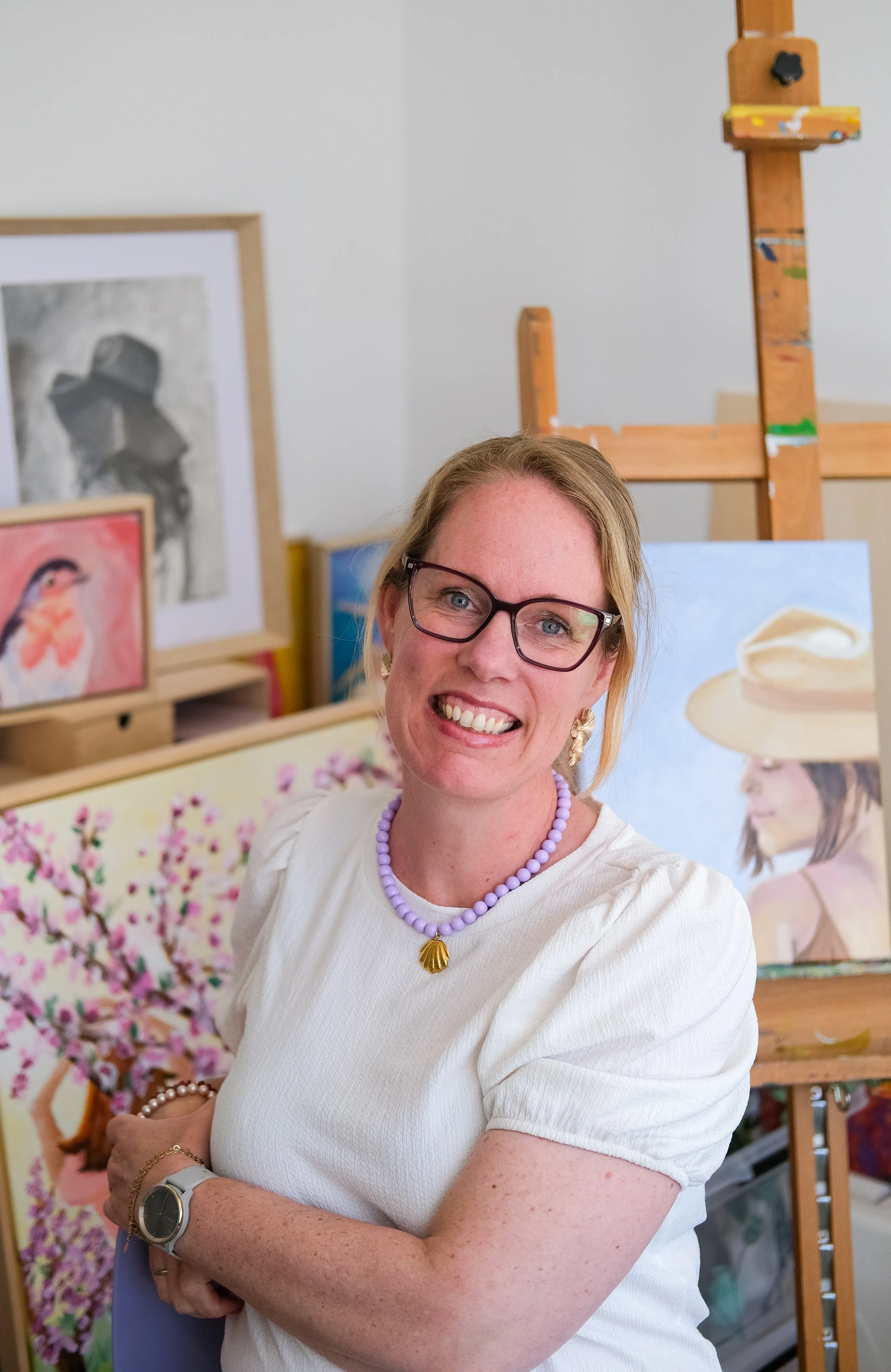 Vrouw met bril, paarse ketting en smartwatch, lachend, in een kunststudio met schilderijen en een houten ezel op de achtergrond.