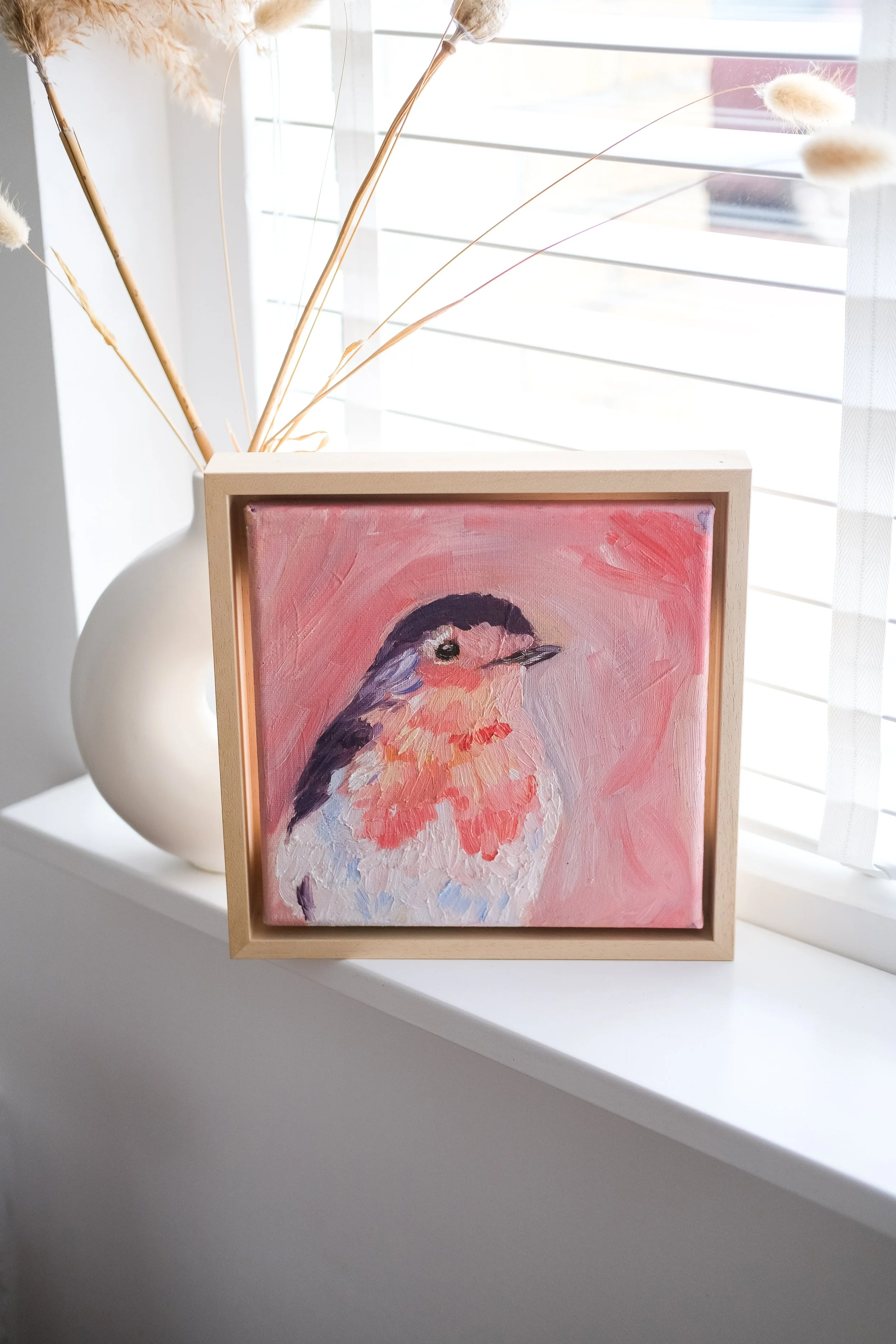 Een schilderij van een vogel met een roze en paarse achtergrond, geplaatst in een houten frame. Het schilderij staat op een wit oppervlak naast een witte vaas met gedroogde planten, bij een raam met horizontale jaloezieën.