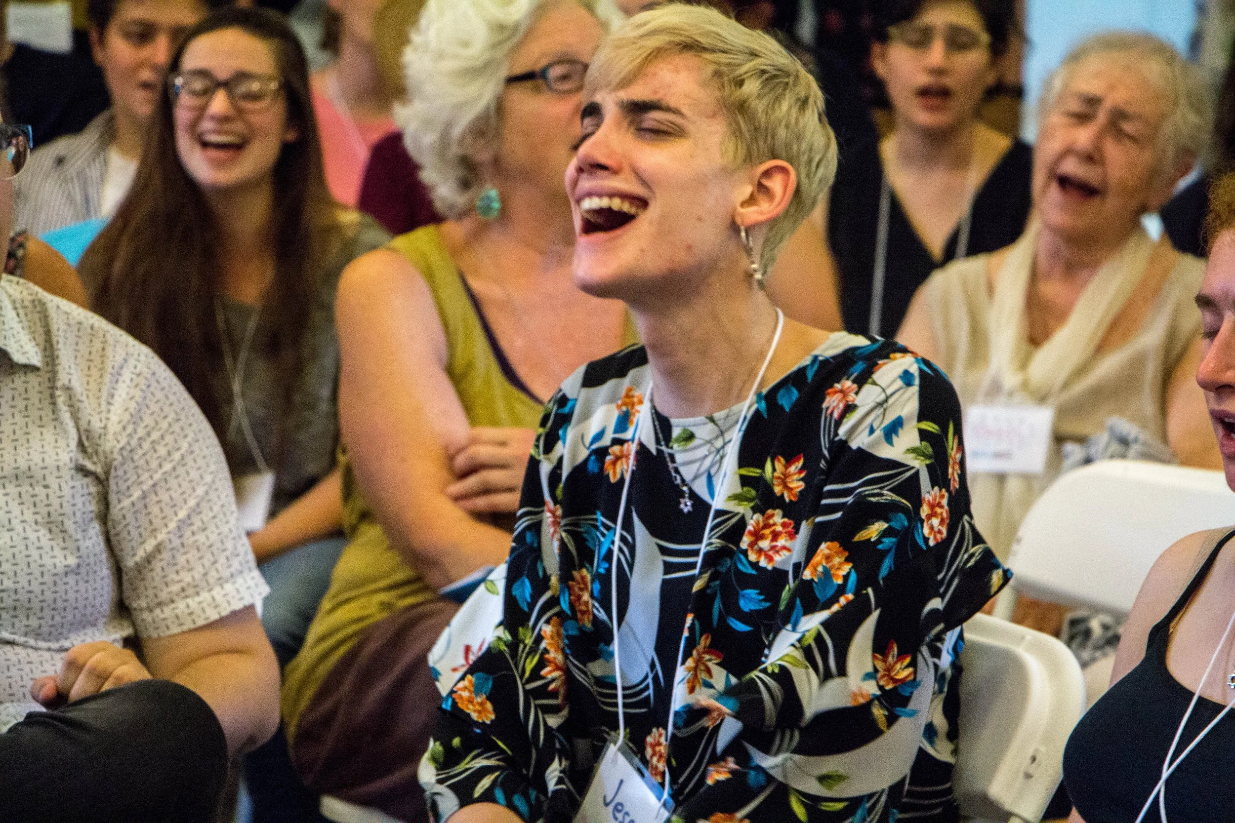A photo of a person with short blond hair, sitting and wearing a name tag, passionately singing, with others singing behind them in the background.