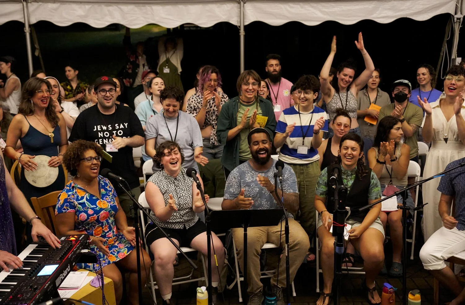 A crowd sits and stands under a large white tent, singing, clapping, and smiling