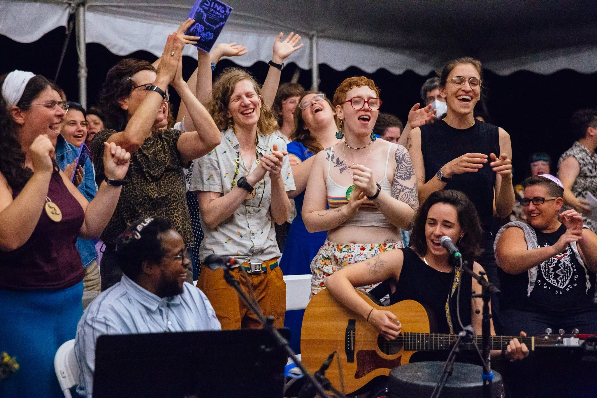 A photo from the 2025 Summer Retreat participatory concert; a group of people sing, some with hands in the air, surrounding teachers playing guitar and singing into mics