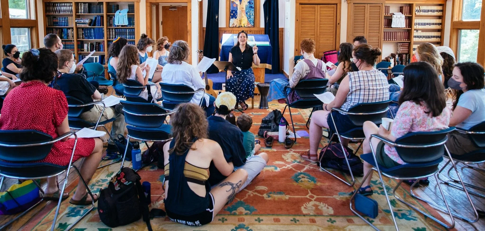 Anat sits at the front of a room, teaching a crowd of singers, reading from paper