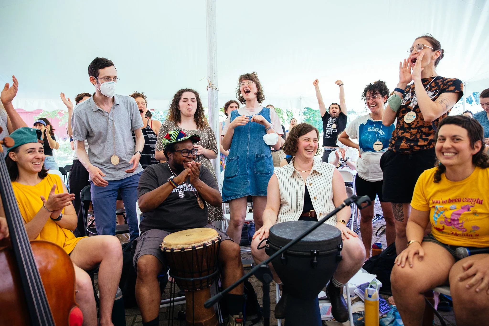 A photo of a crowd sitting and standing under a large white tent, singing loudly and smiling, with drums at the center.