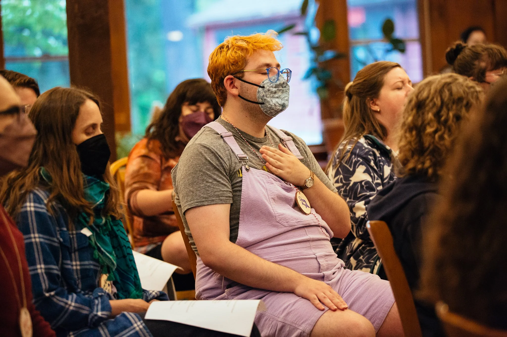 A crowd sits indoors, some wearing masks, singing soulfully. One person puts a hand in their heart and closes their eyes