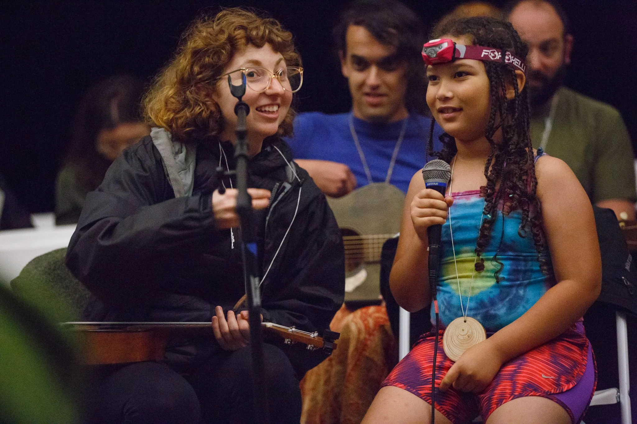 A young adult and a child sit next to each other, the child holding a microphone, and the young adult holding a ukulele
