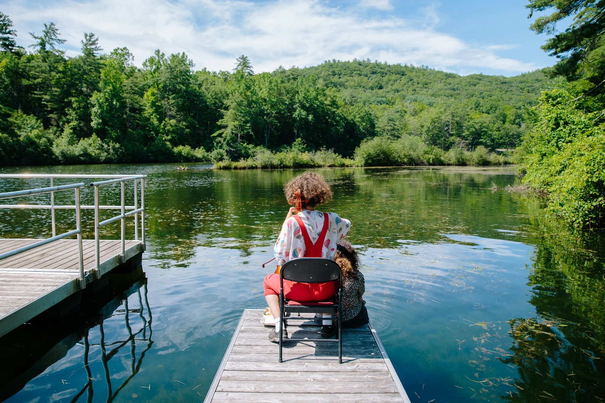 Someone sitting at the edge of a dock in a folding chair, holding an instrument, their back facing the camera, their front facing a calm blue lake surrounded by lush green forest