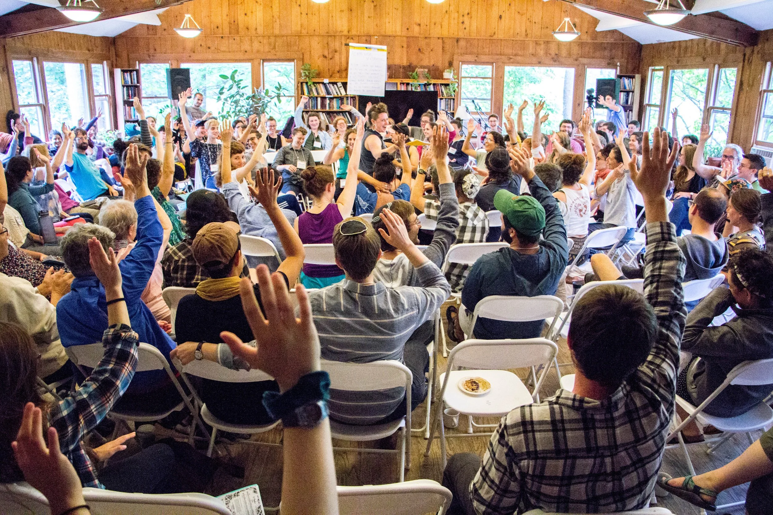 A large crowd sits in concentric circles in a large wooden building, raising their hands in the air