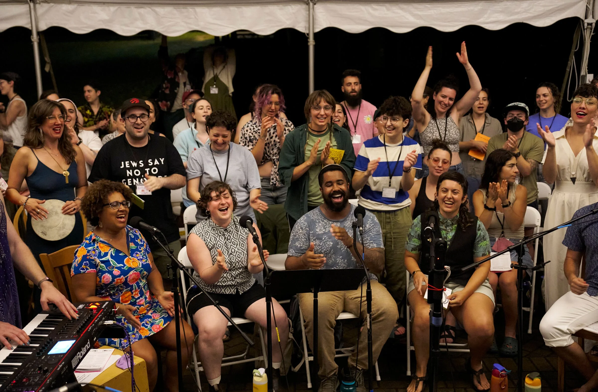 A crowd sits and stands under a large white tent, singing, clapping, and smiling