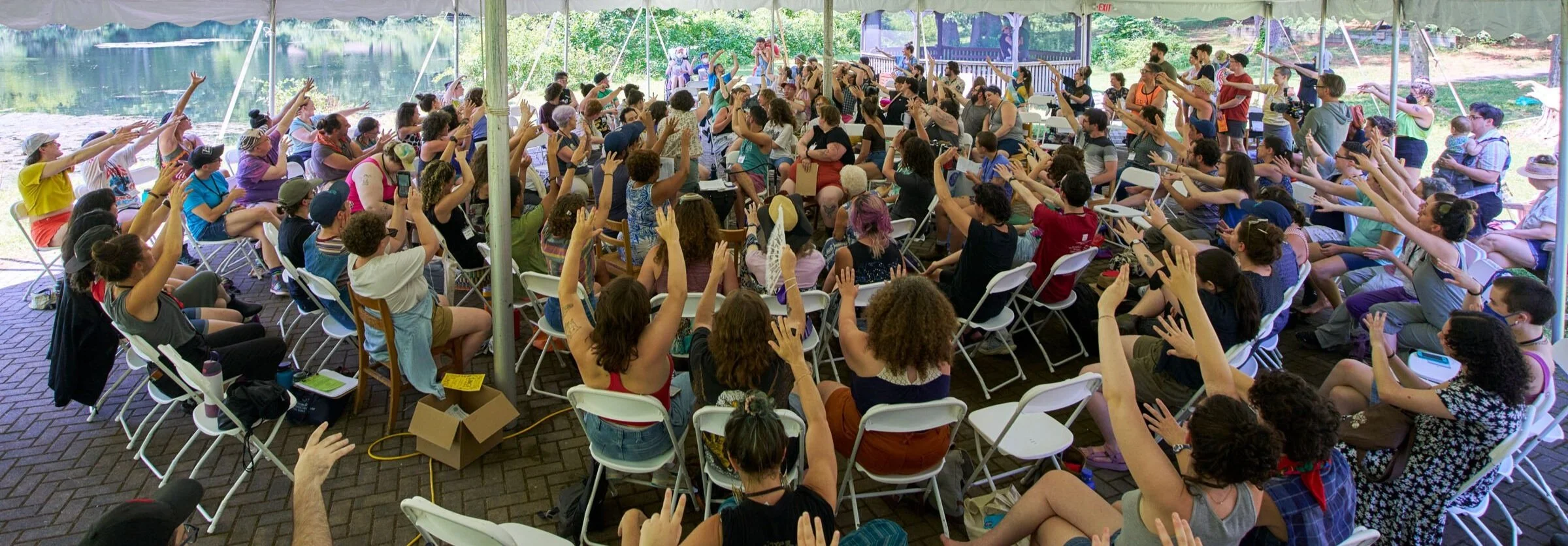 A large group of people outdoors under a large white tent, sitting in folding chairs in concentric circles, with arms up and pointed to the center of the circle