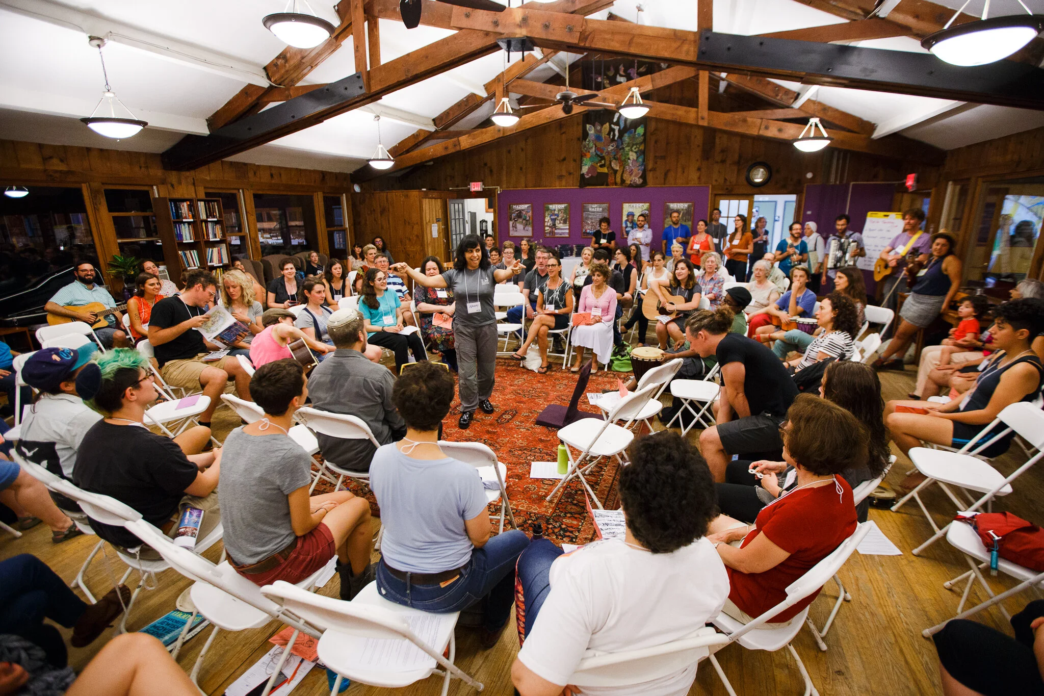 A large crowd sits and sings in folding chairs in a circle, with one person standing and leading in the middle.