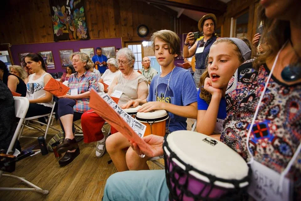 Two kids sing off a sheet of paper, one playing the drums. A crowd of adults sit behind them, singing along as well.