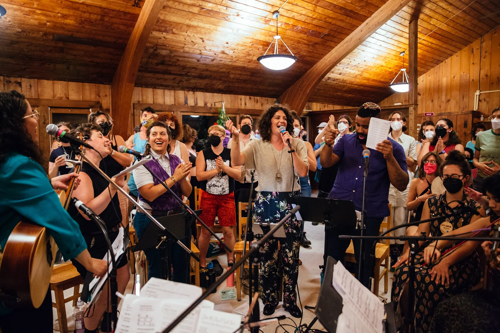 A crowd of people singing, mostly standing, in a large room with wood detailing. Some hold instruments or are singing into a microphone