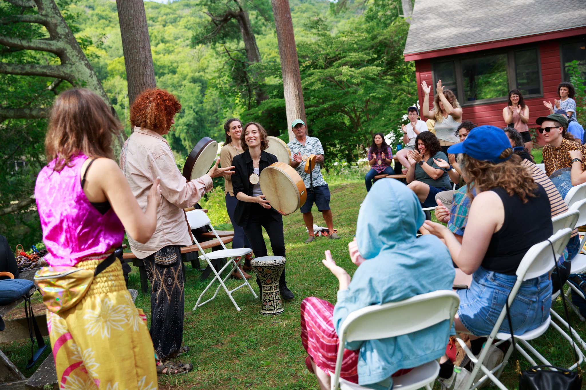 A group of people outside on a sunny day, some sitting in folding chairs and some standing. At the front, people stand with handheld drums