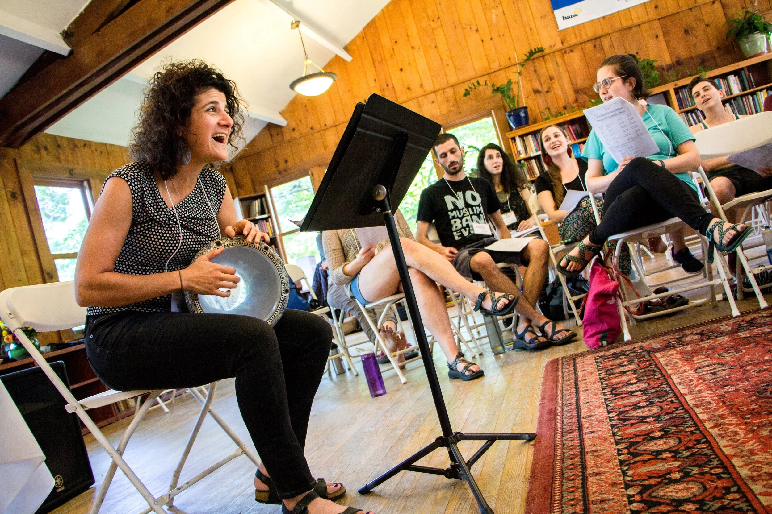 Galeet leading a room in song, with a group sitting behind her. She holds a drum and reads off a music stand.