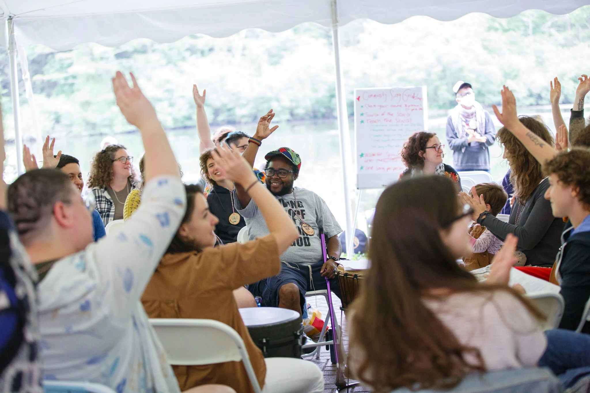A photo of a crowd sitting in folding chairs under a large white tent, raising their hands and smiling