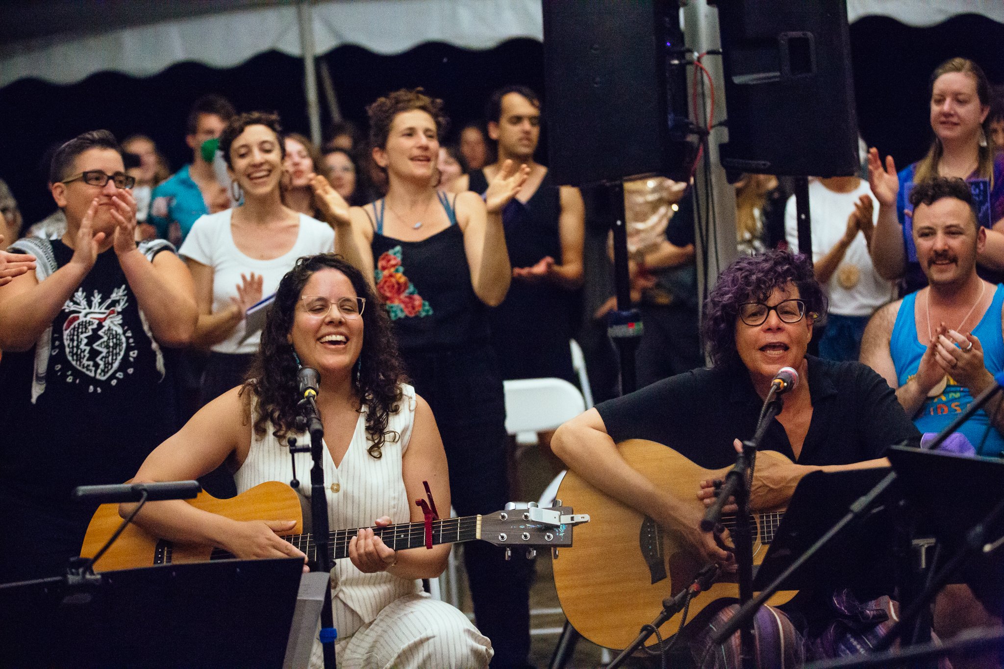 Two people sit in front of a crowd, playing guitar and sinigng. Behind them is a crowd mostly on their feet, clapping and singing