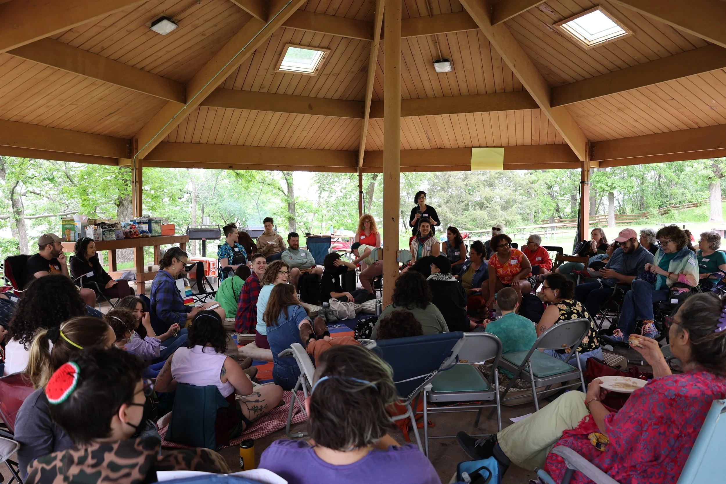 A photo of a crowd gathered under a large wooden awning, sitting in concentric circles