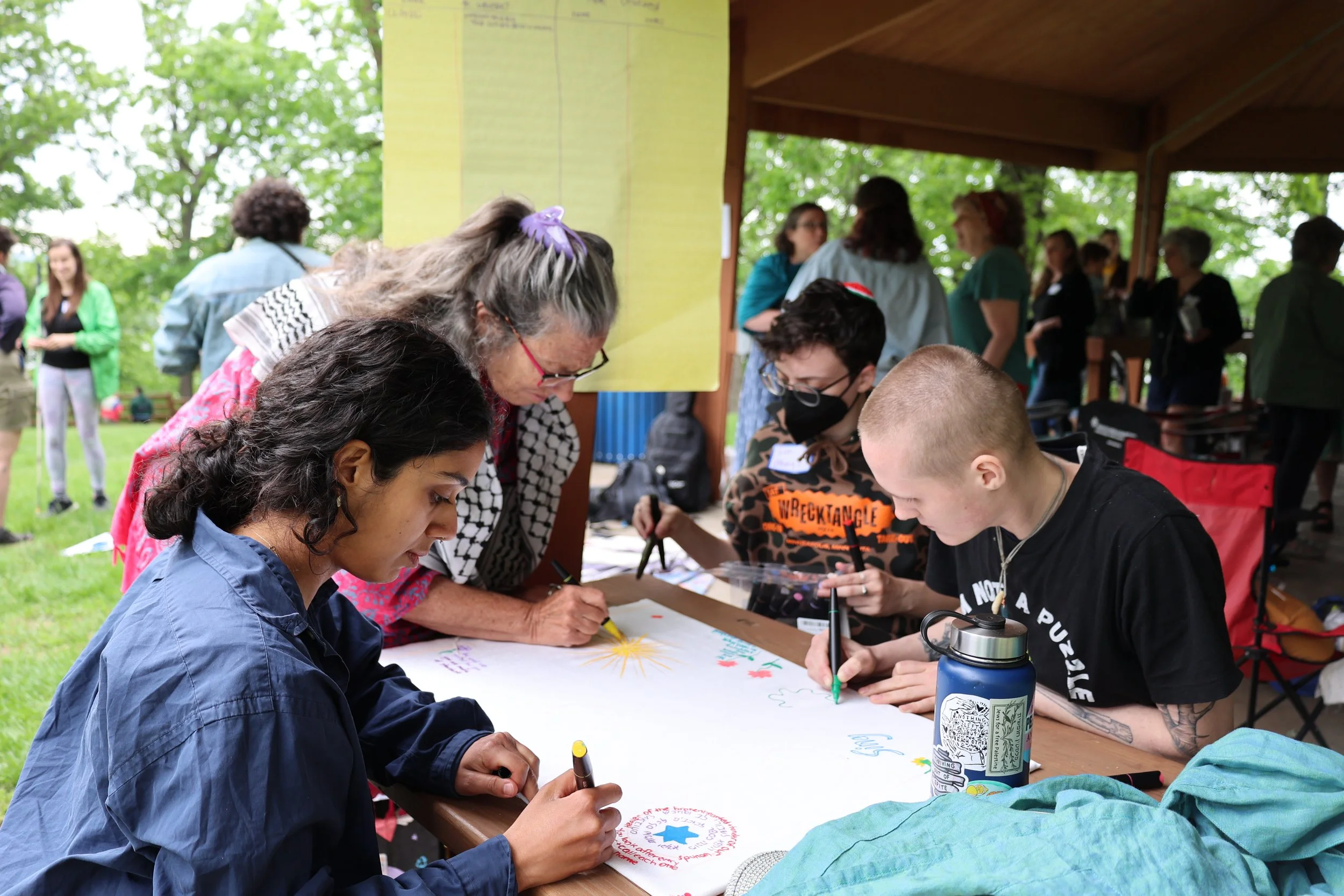 4 people gather around a table outdoors, drawing on a large sheet of butcher paper together