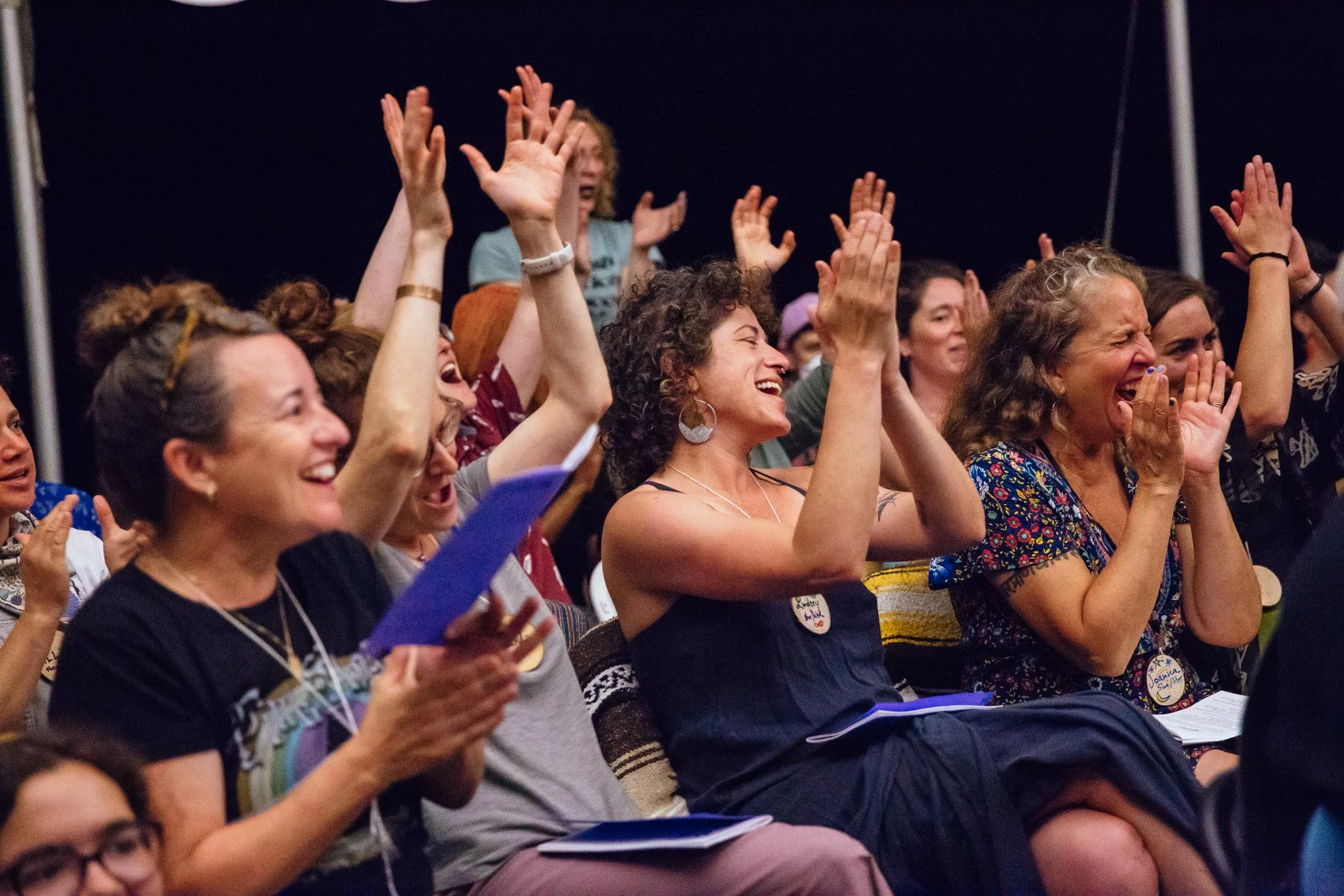 A photo of a crowd sitting and singing and clapping their hands or raising their hands in the air