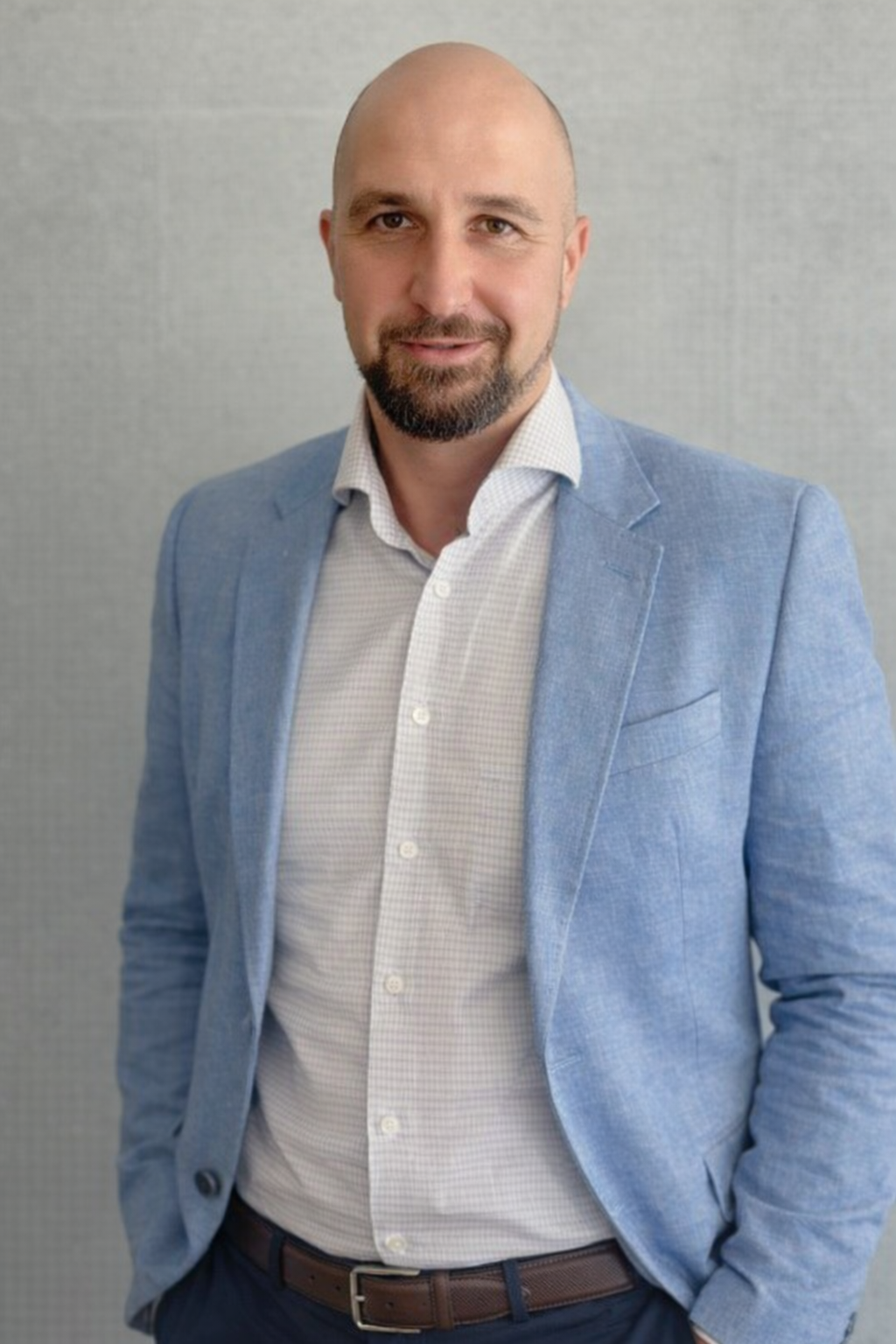A man with a beard and shaved head wearing a light blue blazer, white dress shirt, and dark trousers stands against a plain light grey background.