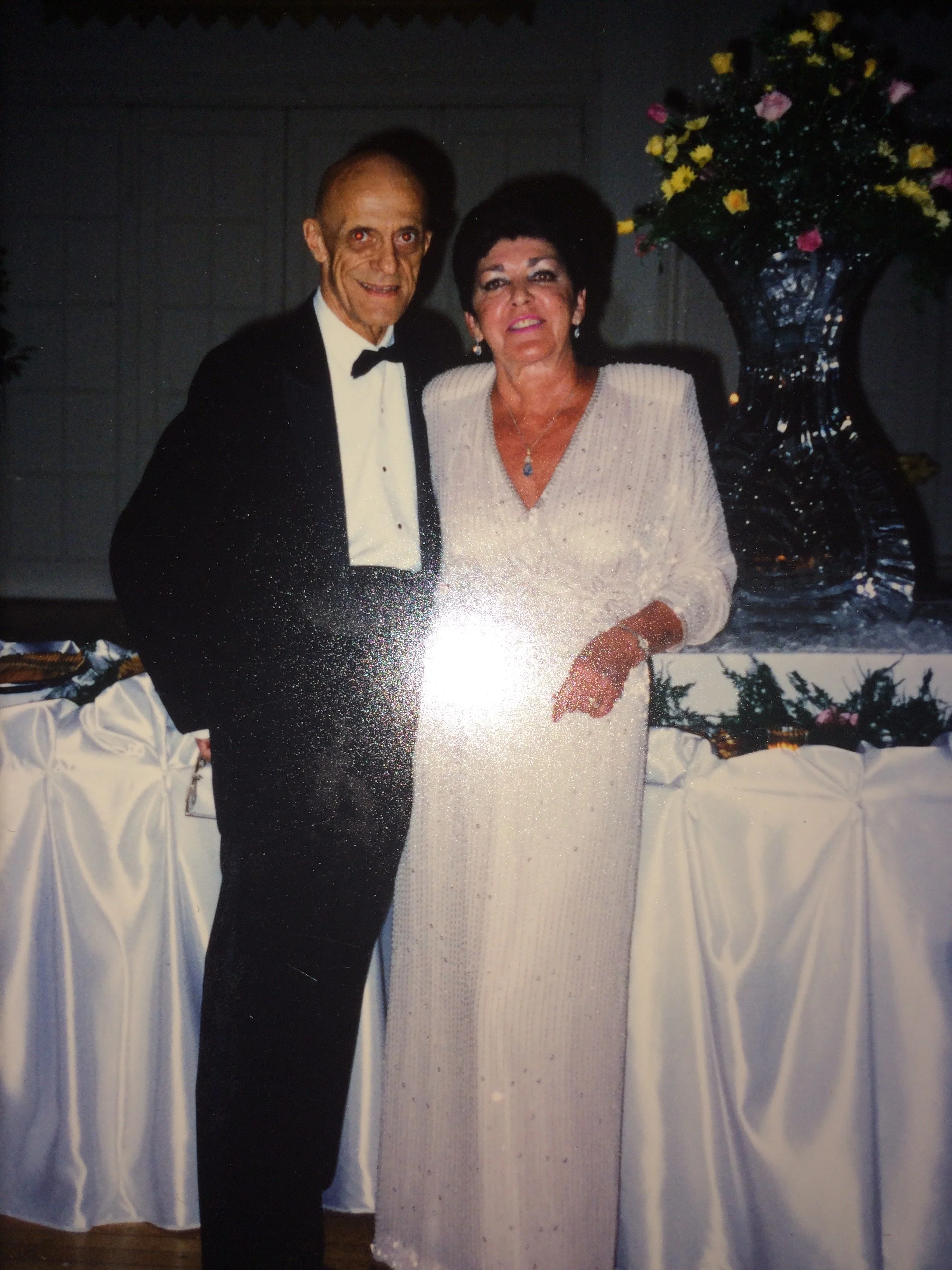 An elderly couple dressed in formal attire with the man in a black tuxedo and the woman in a white, beaded gown, standing together at a celebration next to a large floral arrangement in a dark corner.