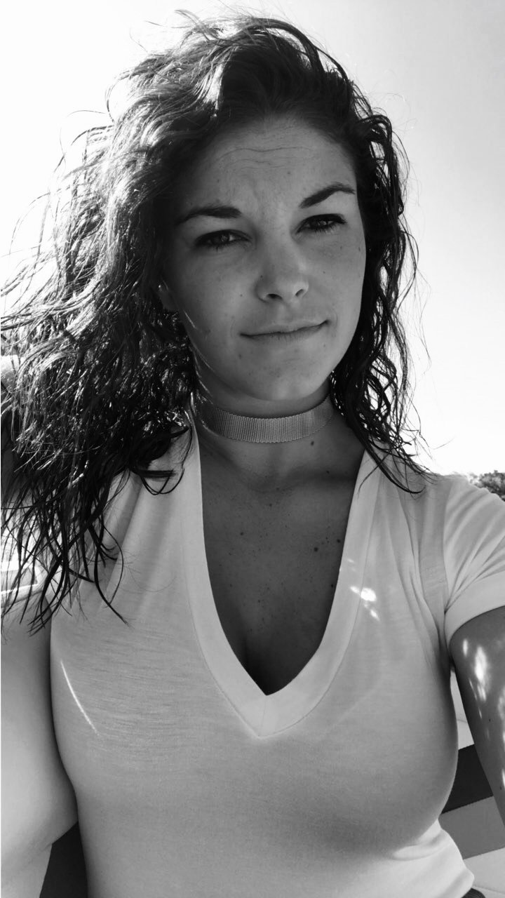 Black and white photo of a young woman with curly hair, wearing a choker necklace and a v-neck t-shirt, outdoors on a sunny day.