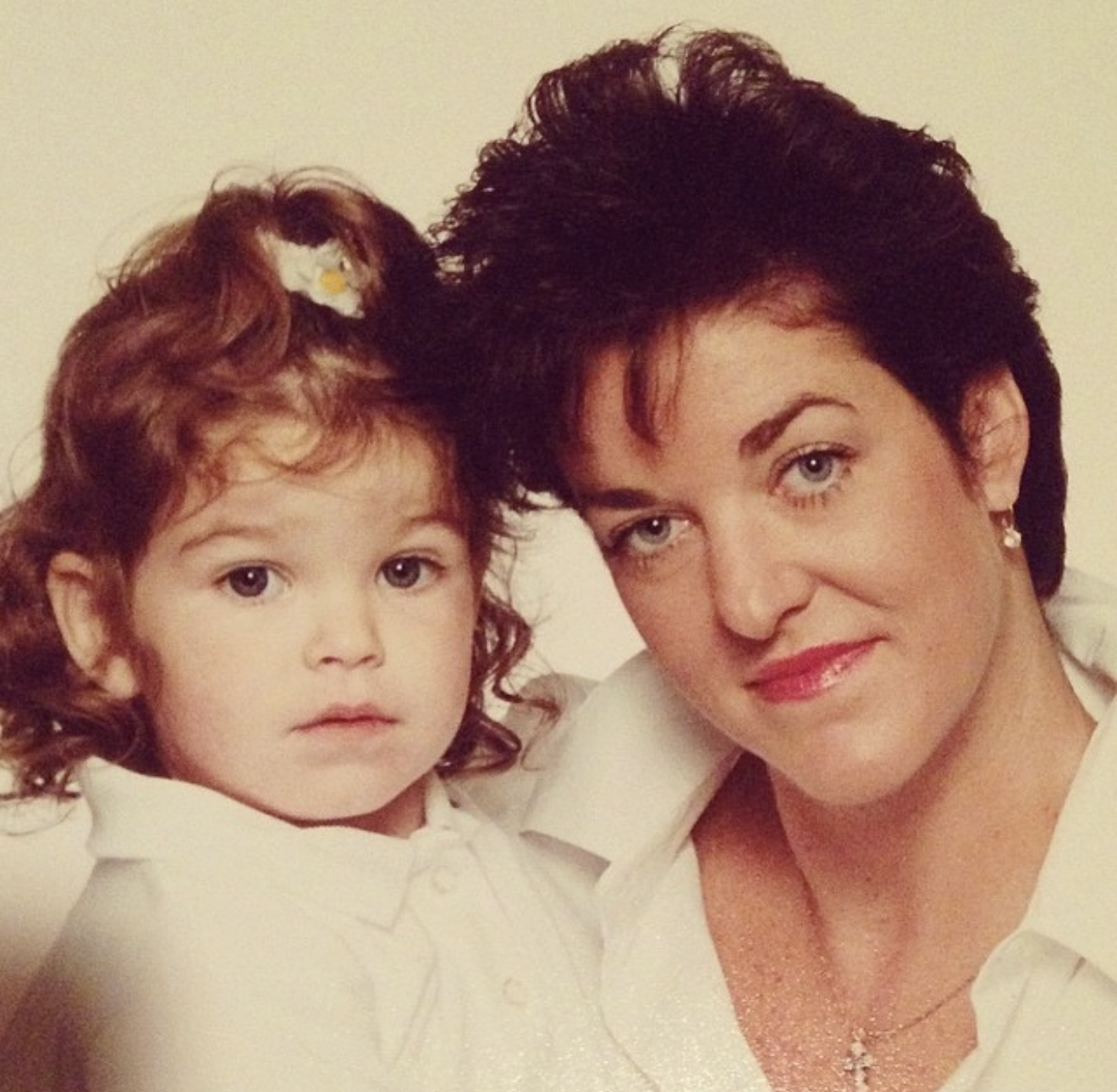 A woman and a young girl with curly red hair and fair skin are close together, both wearing white shirts. The woman has short, dark hair, and the girl has a hair bow and looks directly at the camera. The woman is smiling slightly, with makeup and jewelry visible.