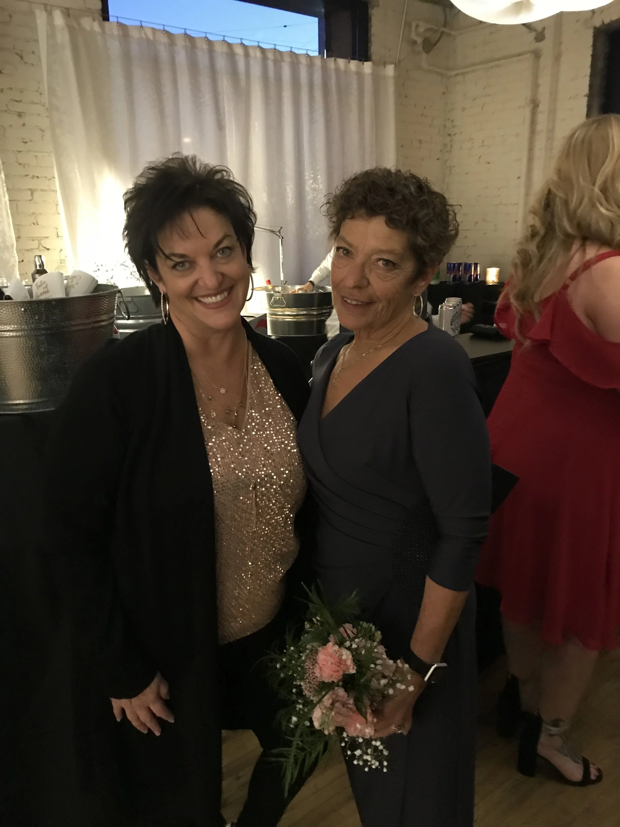 Two women, one with short dark hair and the other with curly gray hair, dressed in formal attire, posing at an indoor event. The woman with short dark hair is smiling and wearing a black jacket and a sequined top. The woman with curly gray hair is holding a bouquet of pink and white flowers, and is wearing a dark dress. There are other people and tables with drinks in the background.