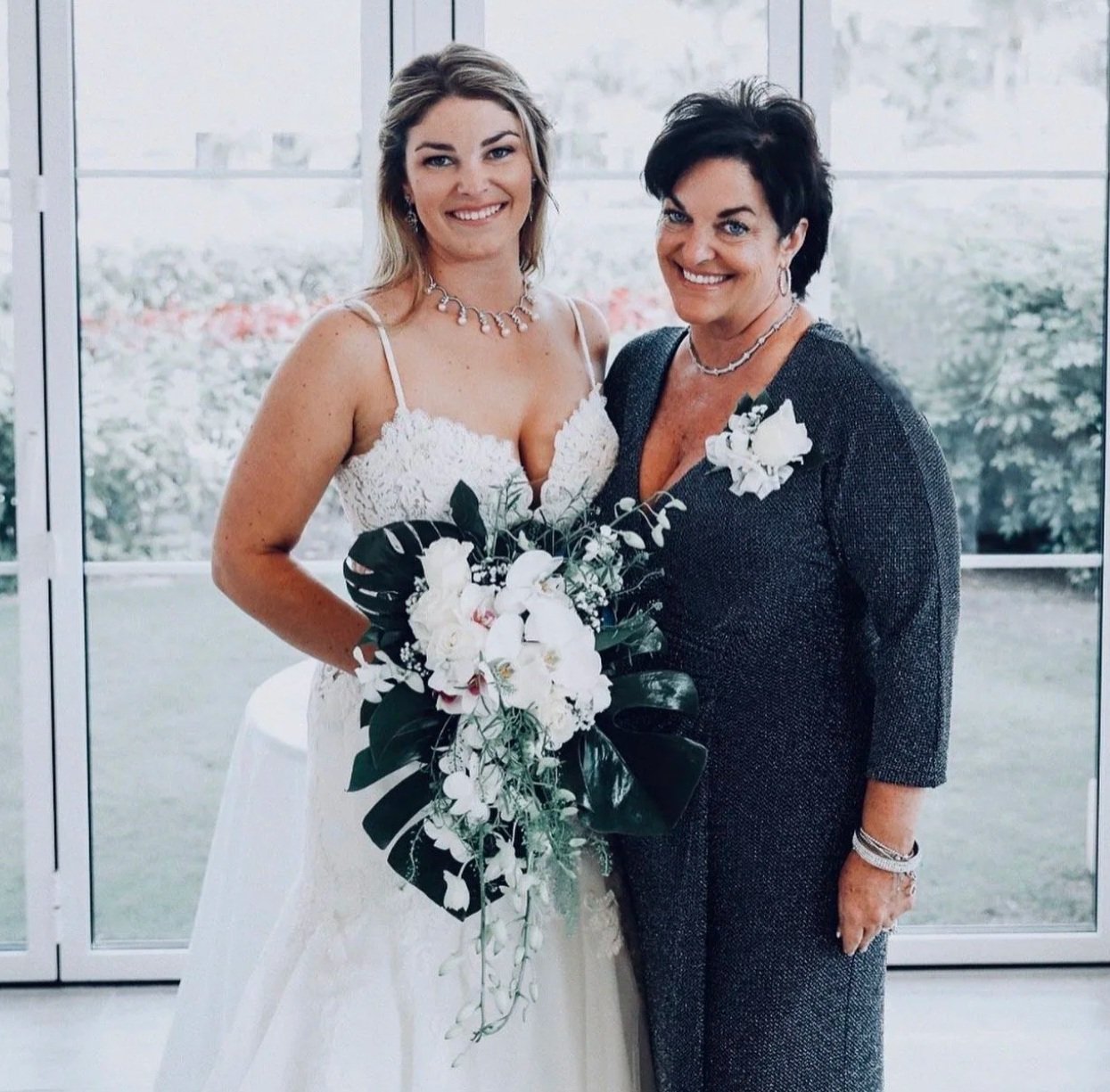 Two women in formal attire, one in a wedding dress holding a bouquet, standing indoors in front of large windows with garden view.
