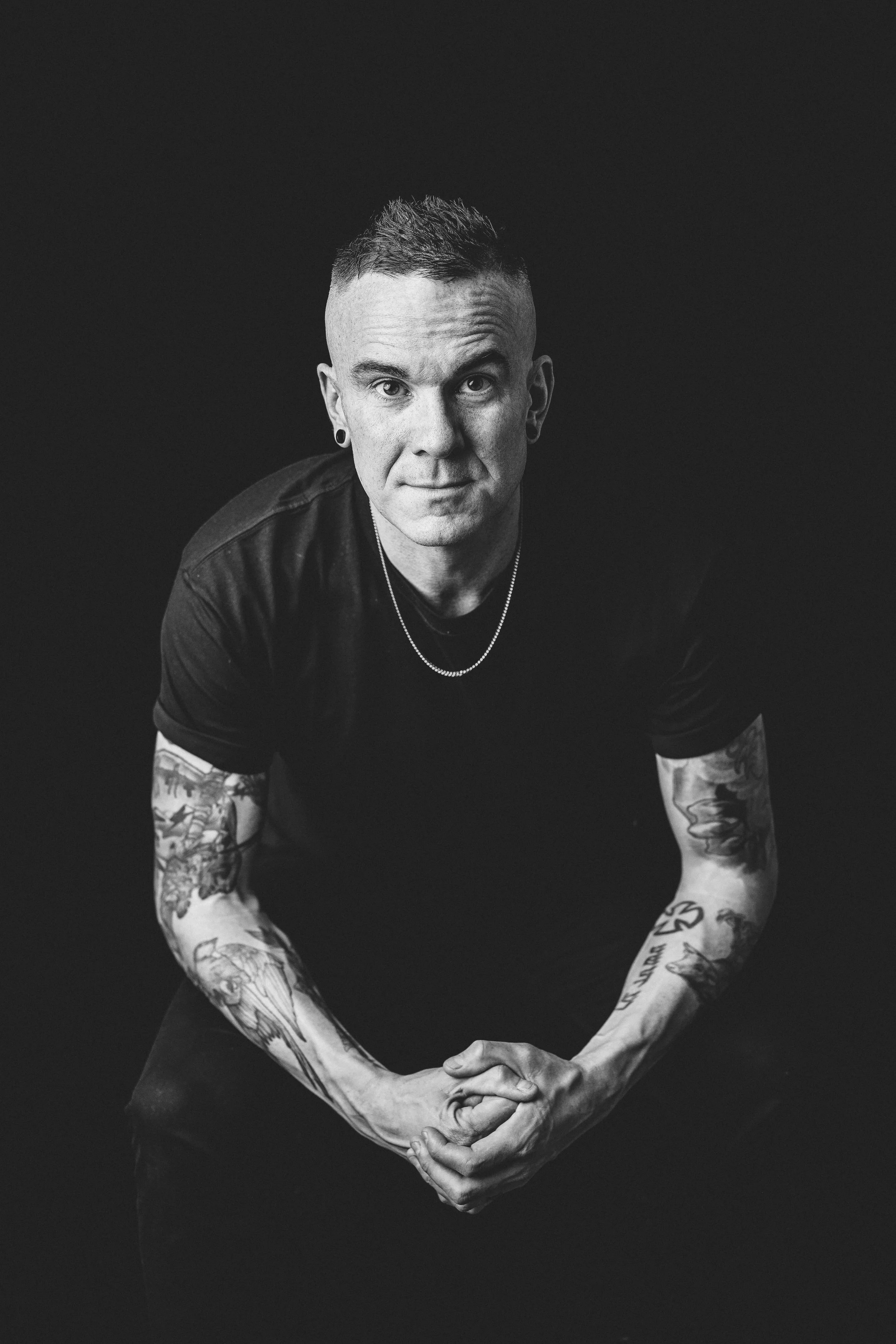 Black and white portrait of a man with tattoos on his arms, wearing a black shirt, a necklace, and earrings, sitting and looking directly at the camera with a neutral expression.