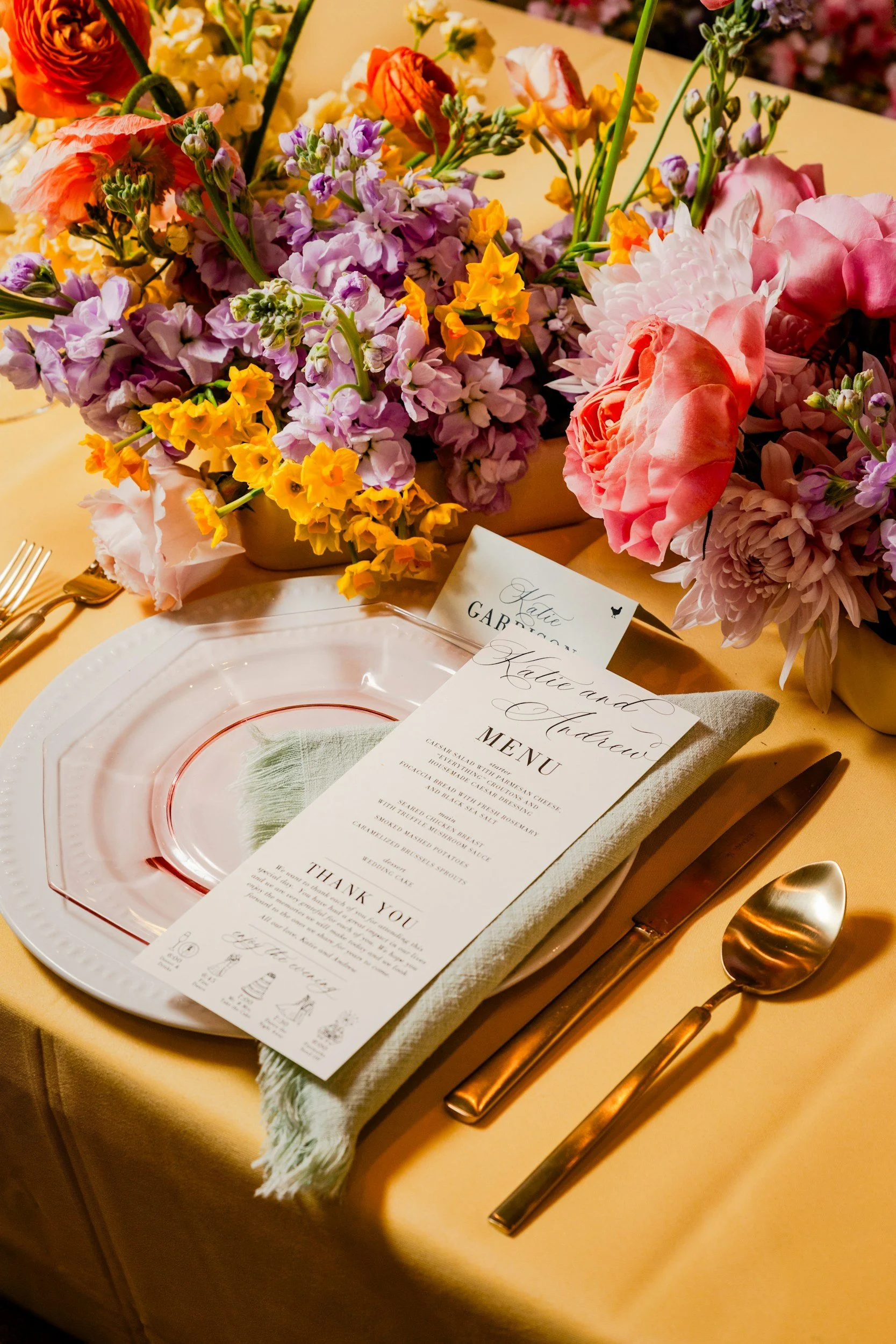 A wedding reception table setting with a floral centerpiece of pink, purple, yellow, and orange flowers, a printed menu, and gold utensils on a beige tablecloth.