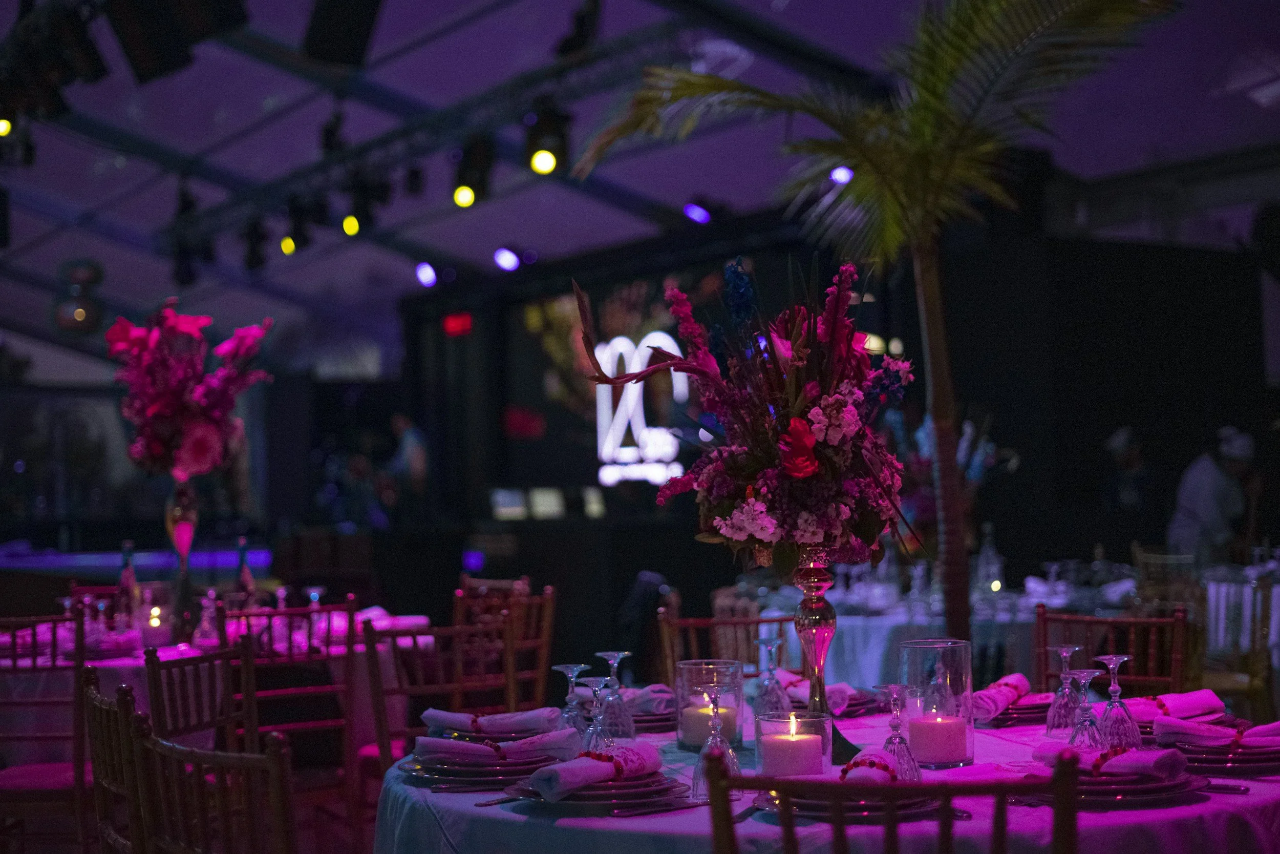 Elegant event setup with round table decorated with pink floral centerpieces, candles, and glassware, in a dimly lit venue with purple and yellow stage lighting.