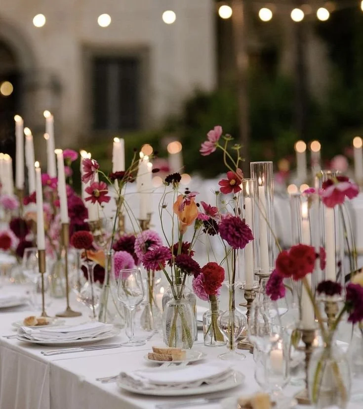 Elegant outdoor dining table decorated with pink and red flowers, tall white candles, and string lights in the background.