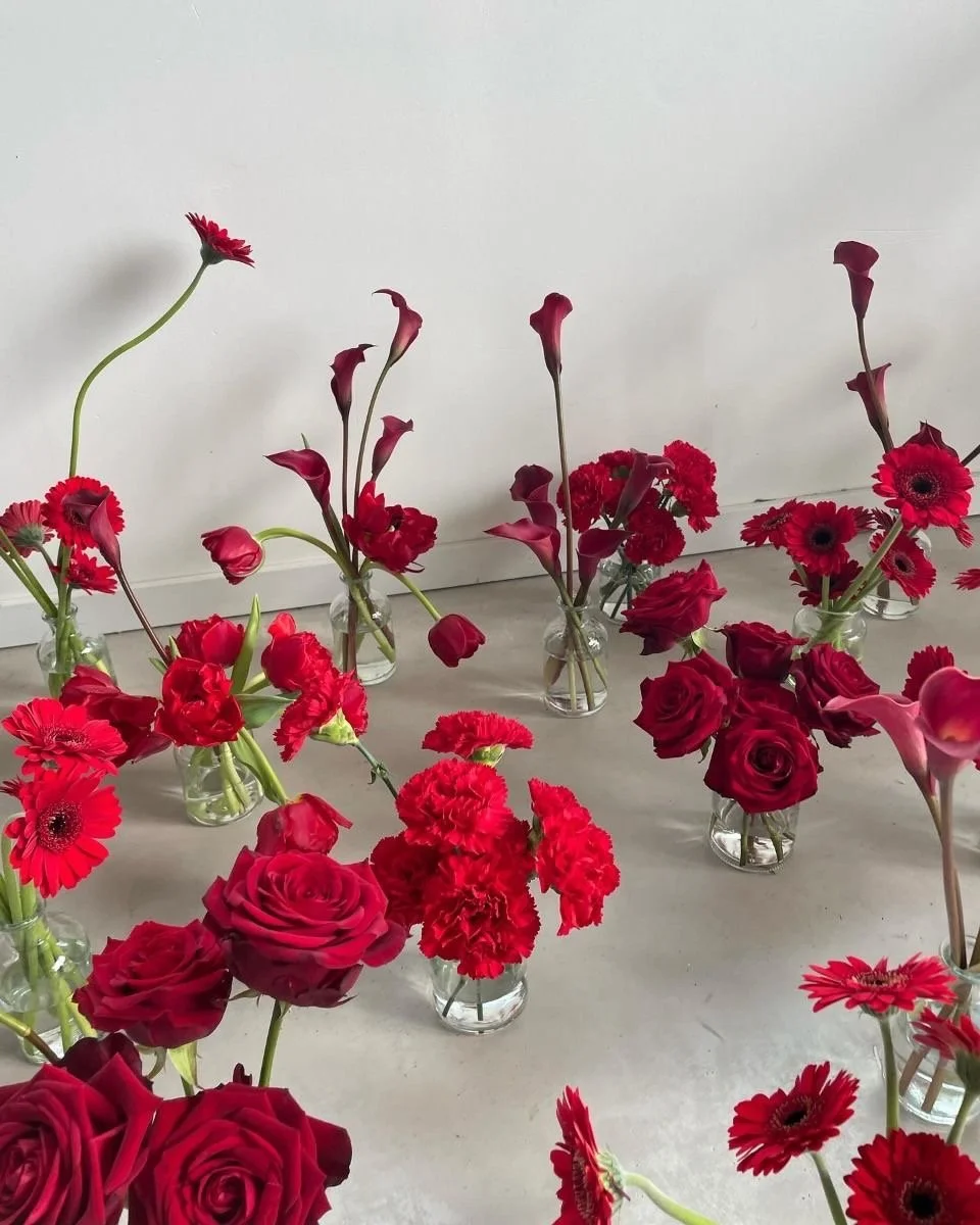 Multiple vases of red and pink flowers arranged on a white floor against a plain white wall.