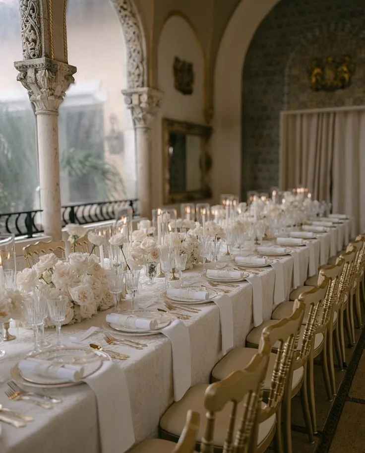 Elegant wedding reception table with white floral centerpieces, candles, and fine china, set in a decorated historic building with arched windows and ornate columns.