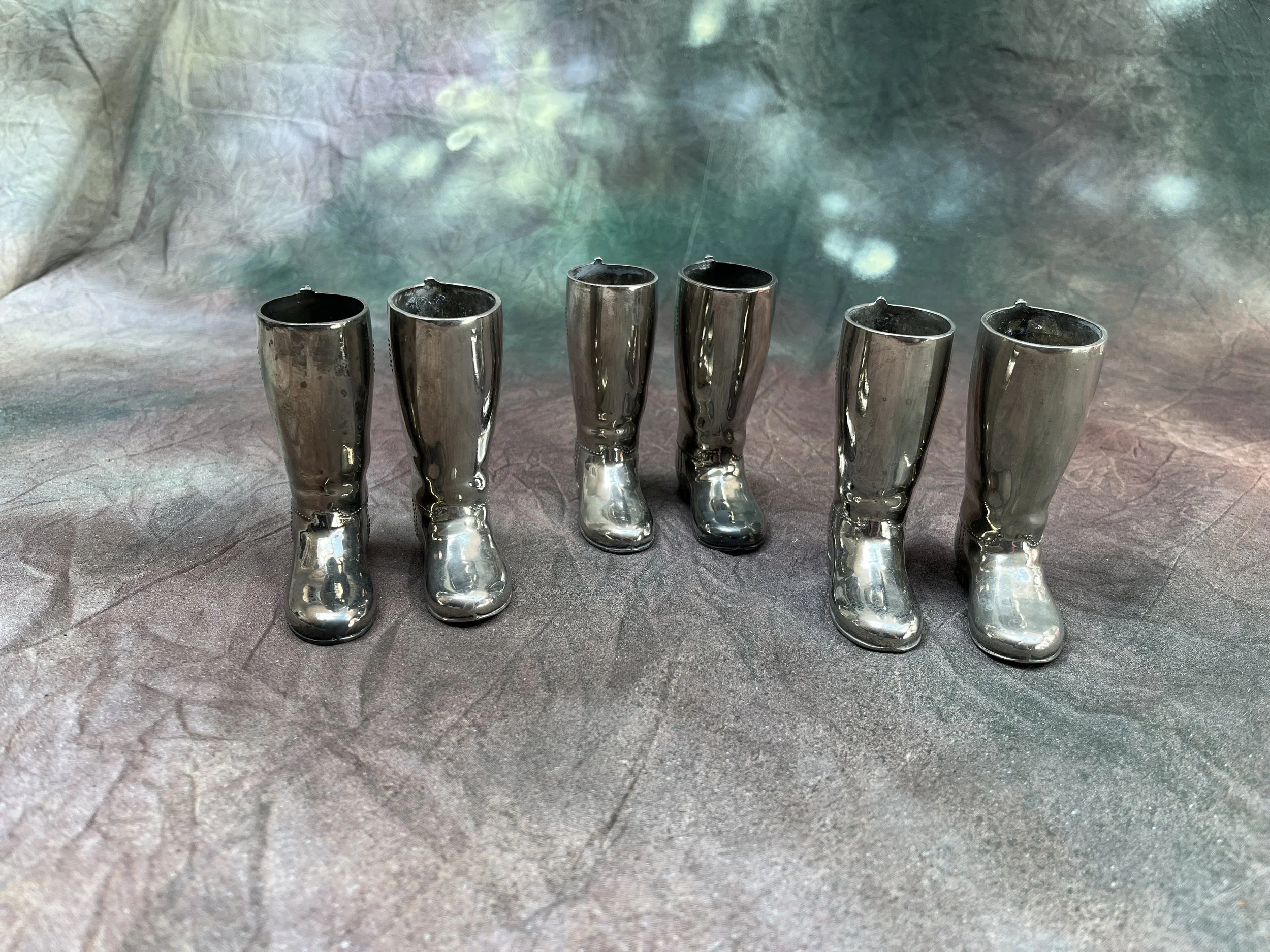 Six shiny silver toy boots are lined up on a textured surface, with a background of an abstract, blurred mixture of colors including green, blue, and pink.