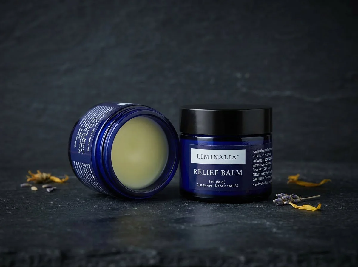 Blue jar of Limnalia Relief Balm with black lid, next to another open jar showing balm inside, on a dark surface with dried lavender and yellow flower petals around.