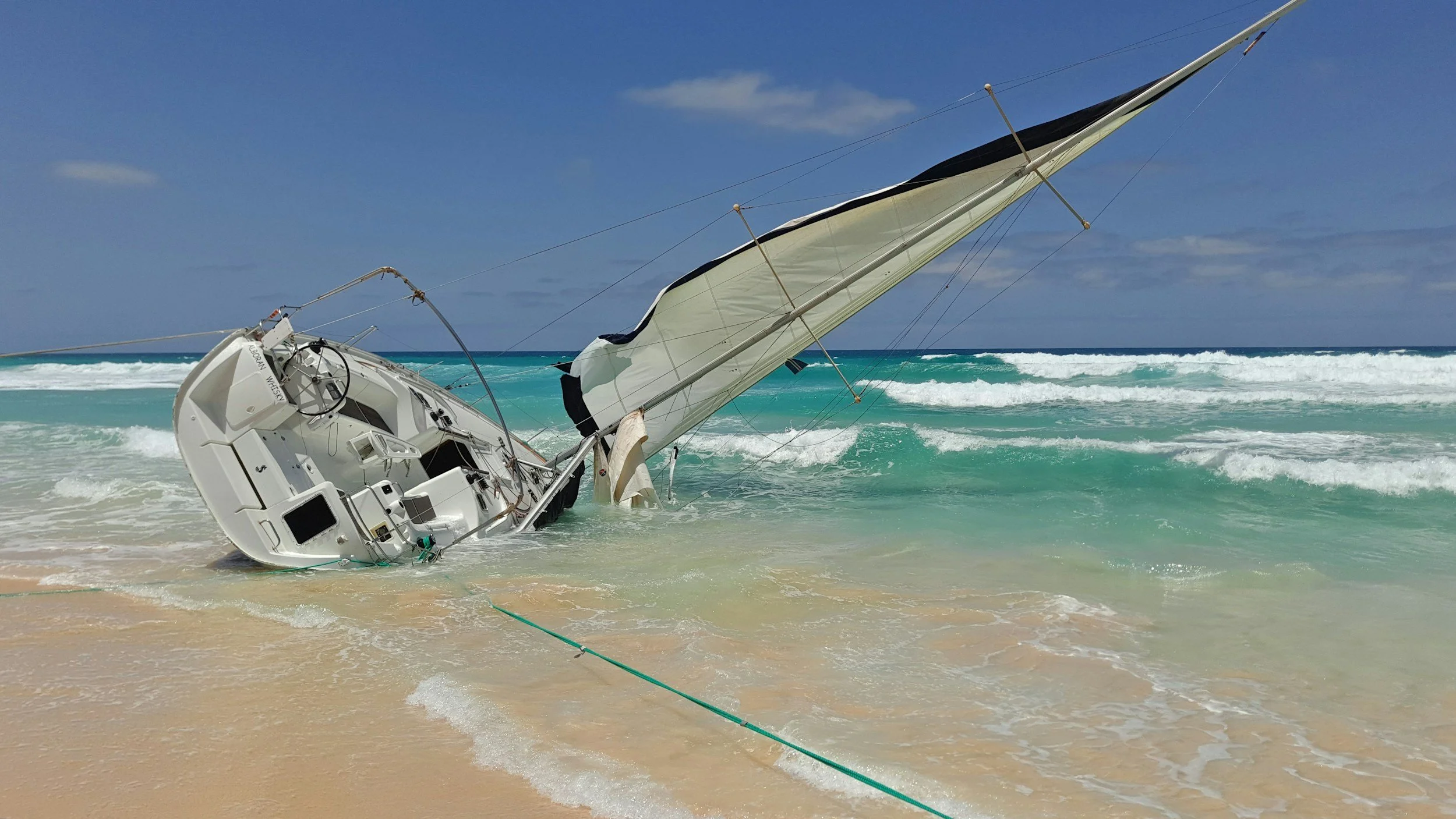 Sailboat capsized on sandy beach, with its mast and sails in the water near turquoise ocean waves under a partly cloudy sky.