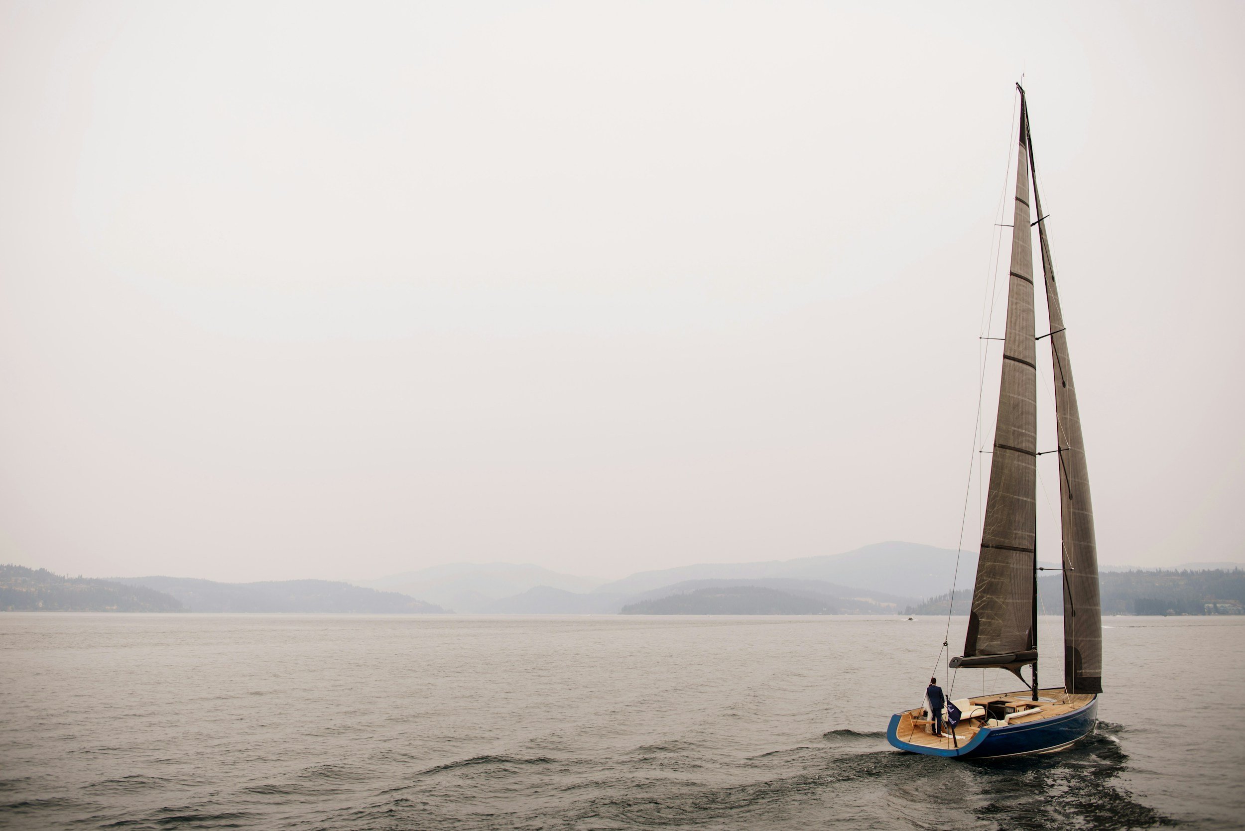 A sailboat with a large sail sailing on a calm body of water, with hills and a faint shoreline in the background, overcast sky