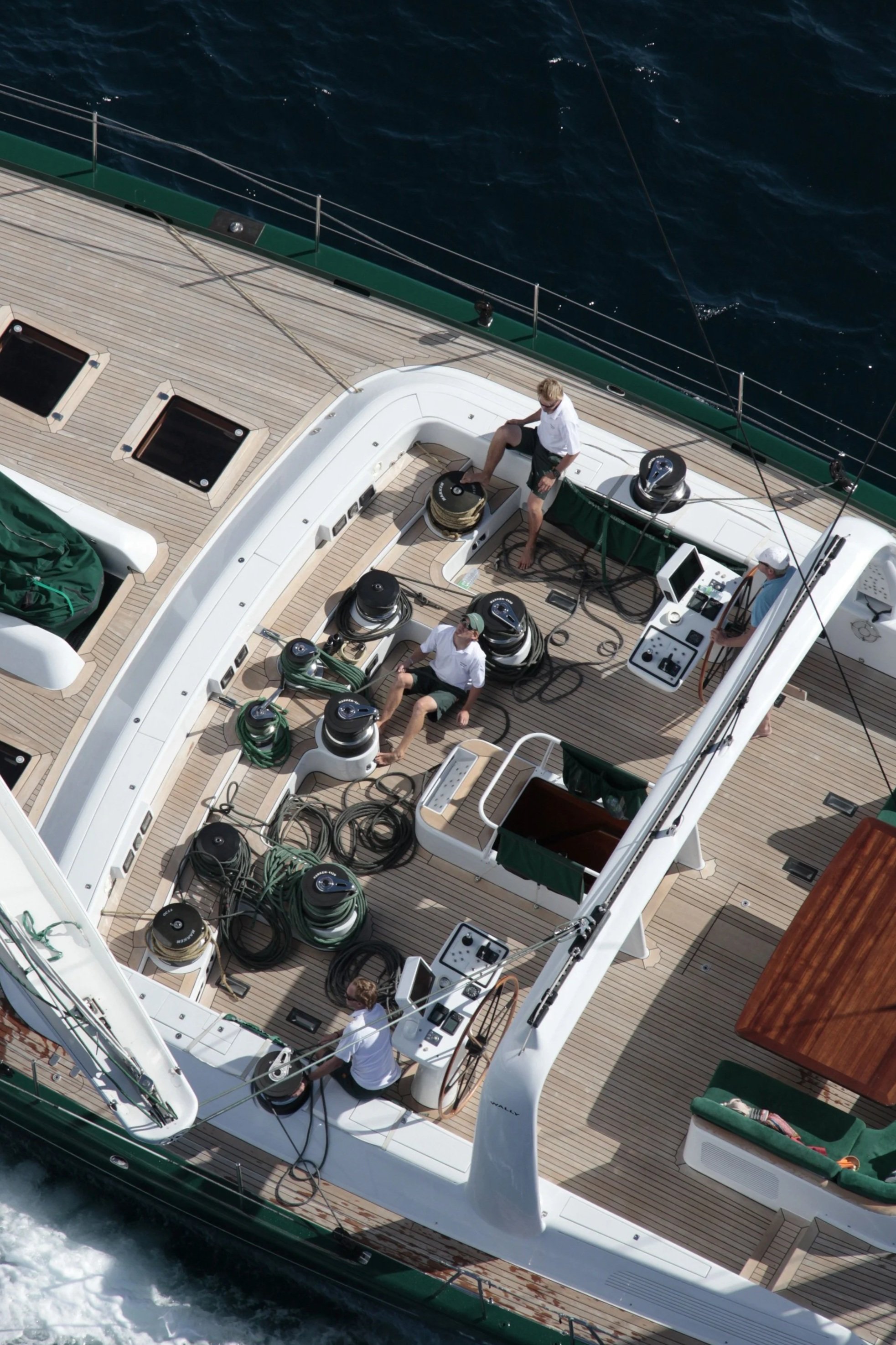 Aerial view of a yacht with crew members working on deck near the ocean. The deck has several winches, ropes, and equipment.