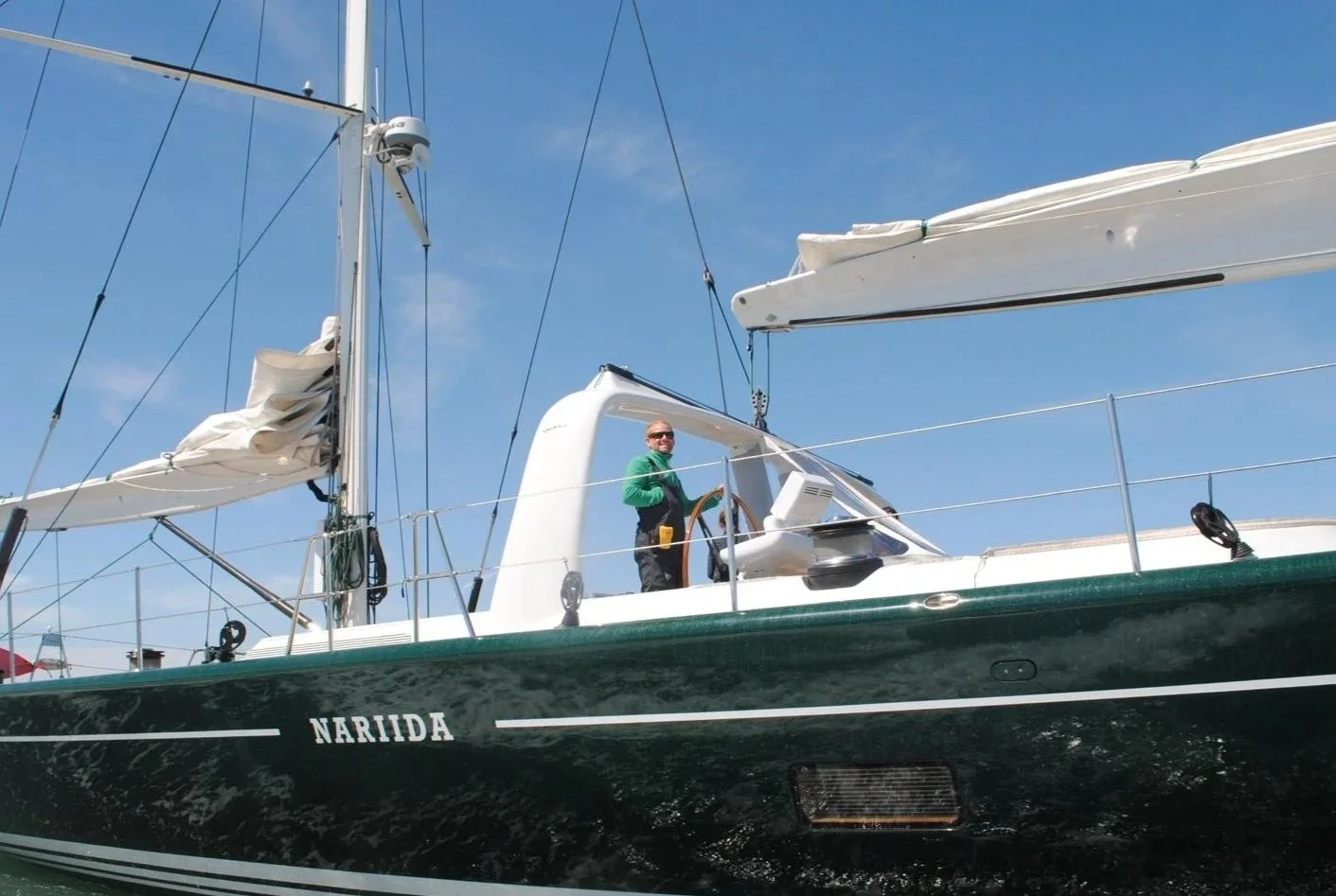 A man standing on a sailboat named Nariida, holding the steering wheel and smiling, with blue sky and clouds in the background.