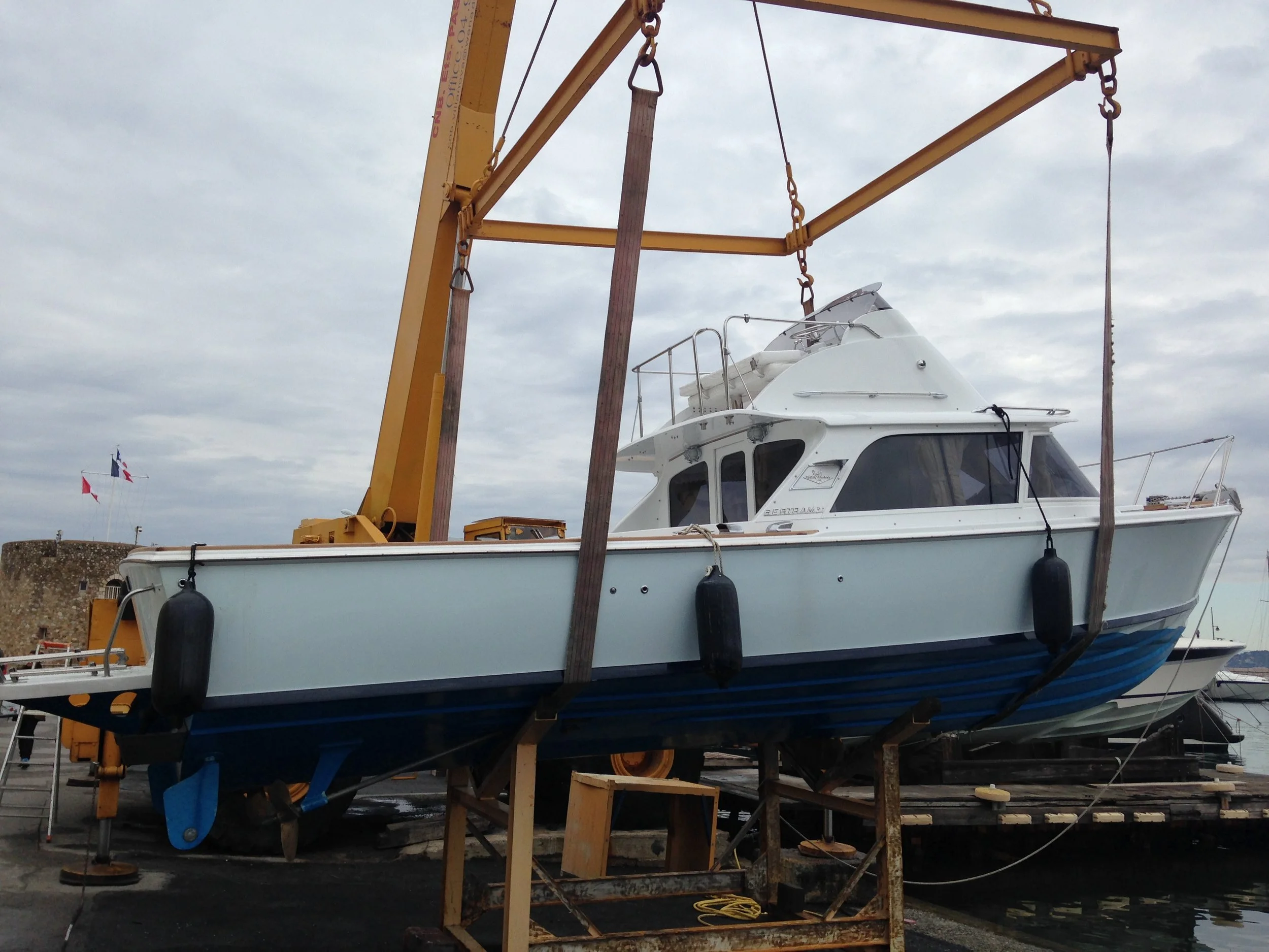 A white boat is suspended on a wooden lift in a boatyard, with the sky and flags in the background.