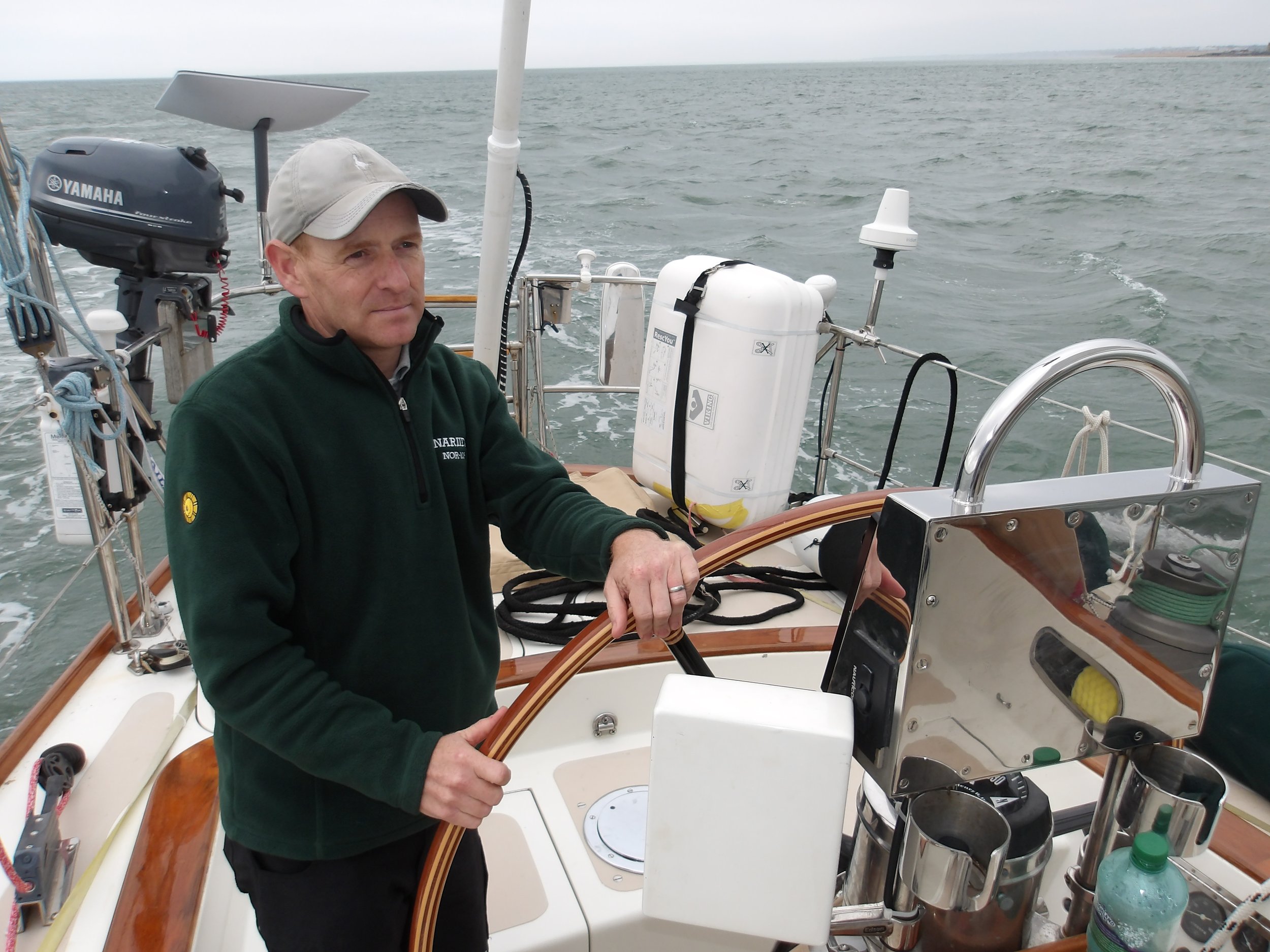 A man wearing a beige cap and a dark green jacket with a logo, standing on a sailboat, steering with a wooden wheel, with the ocean and a distant shoreline in the background.