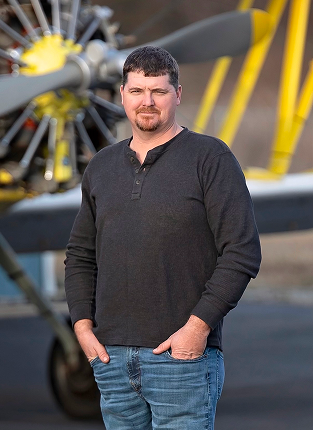Man standing in front of a vintage aircraft, wearing a black sweater and blue jeans.