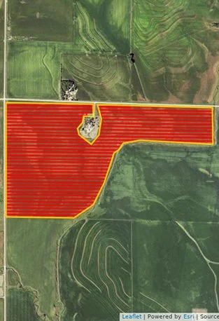 Aerial view of farmland with a large rectangular field highlighted in red, bordered by a yellow line, surrounded by green fields and patches of trees.