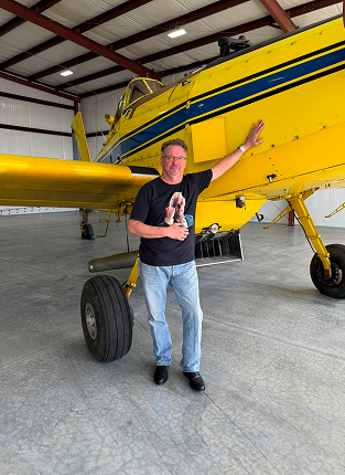 A man standing inside a hangar next to a bright yellow aircraft, holding a small dog, and smiling at the camera.