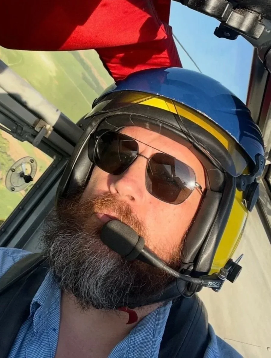 Self-portrait of a man with a beard taking a selfie inside a small aircraft, wearing a blue helmet with a yellow stripe, sunglasses, and a blue checkered shirt.